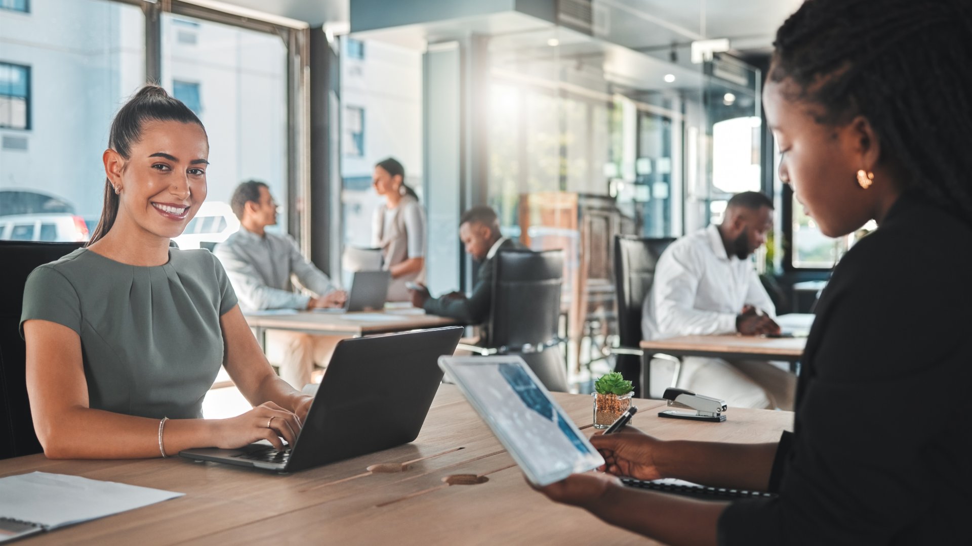 Bright coworking space with people using laptops and tablets around a long table in a glass-walled office.
