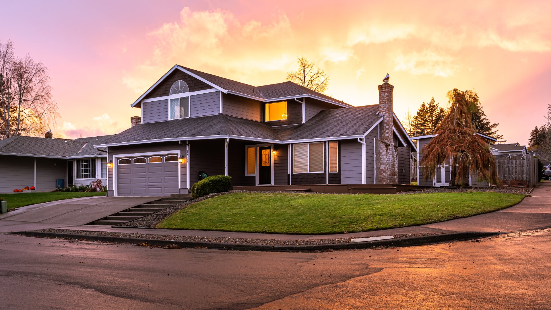 A well-maintained two-story house with a garage and walkway in a neighborhood. The sun is setting behind the house.