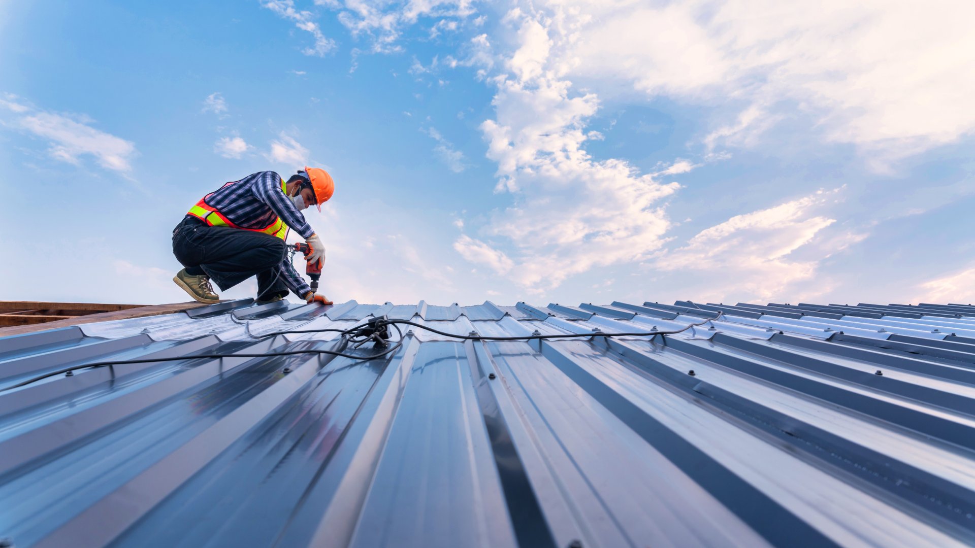 A man in an orange hard hat, safety vest, and gloves uses a nail gun to install sheet metal on a roof.