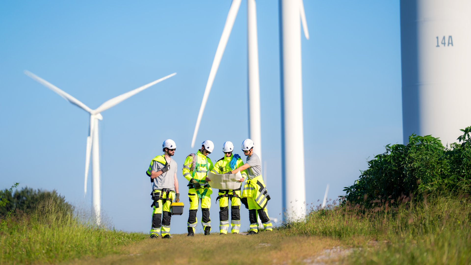  Four people in high-vis work gear and white hard hats standing near a wind turbine and looking at a large piece of paper.