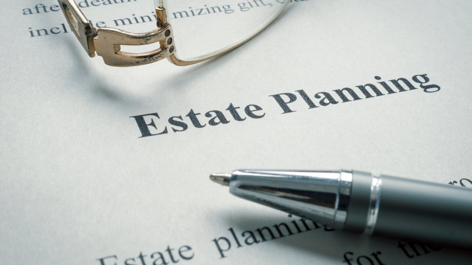 A close-up of a white document that says "Estate Planning" at the top with a pen and eyeglasses on top of it.
