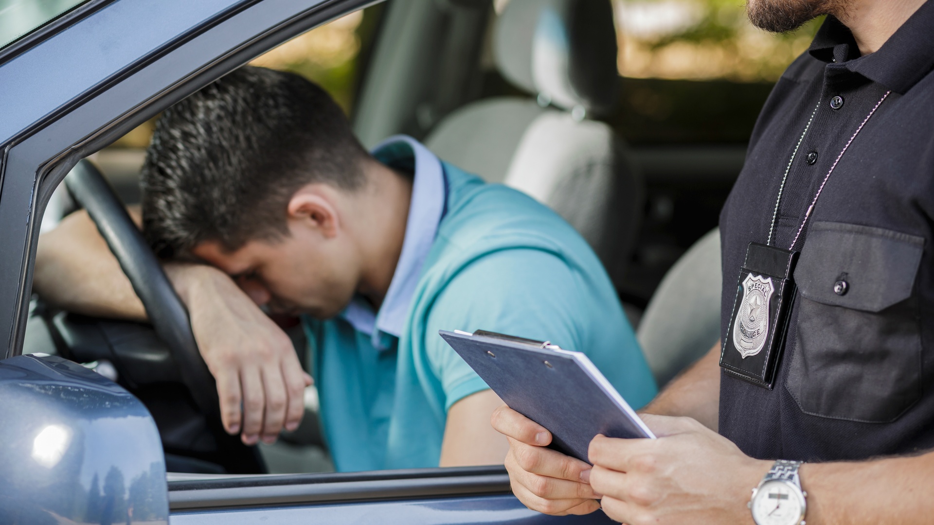 A police officer stands beside a car's open driver's side window. The driver leans with his arm and head on the steering wheel.