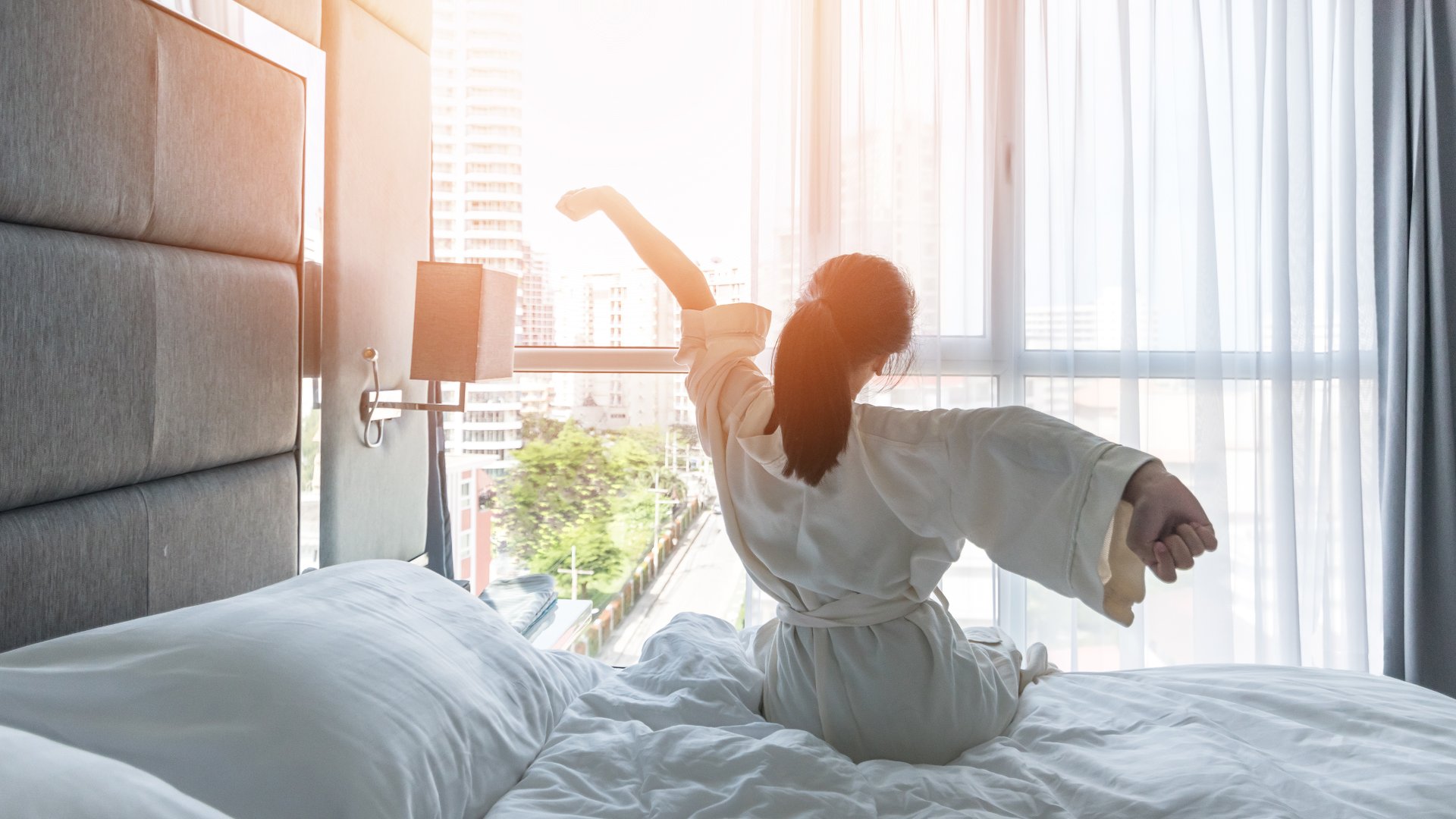 A woman in a white bathrobe is sitting up in bed and stretching her arms as she looks out a window to a city street.