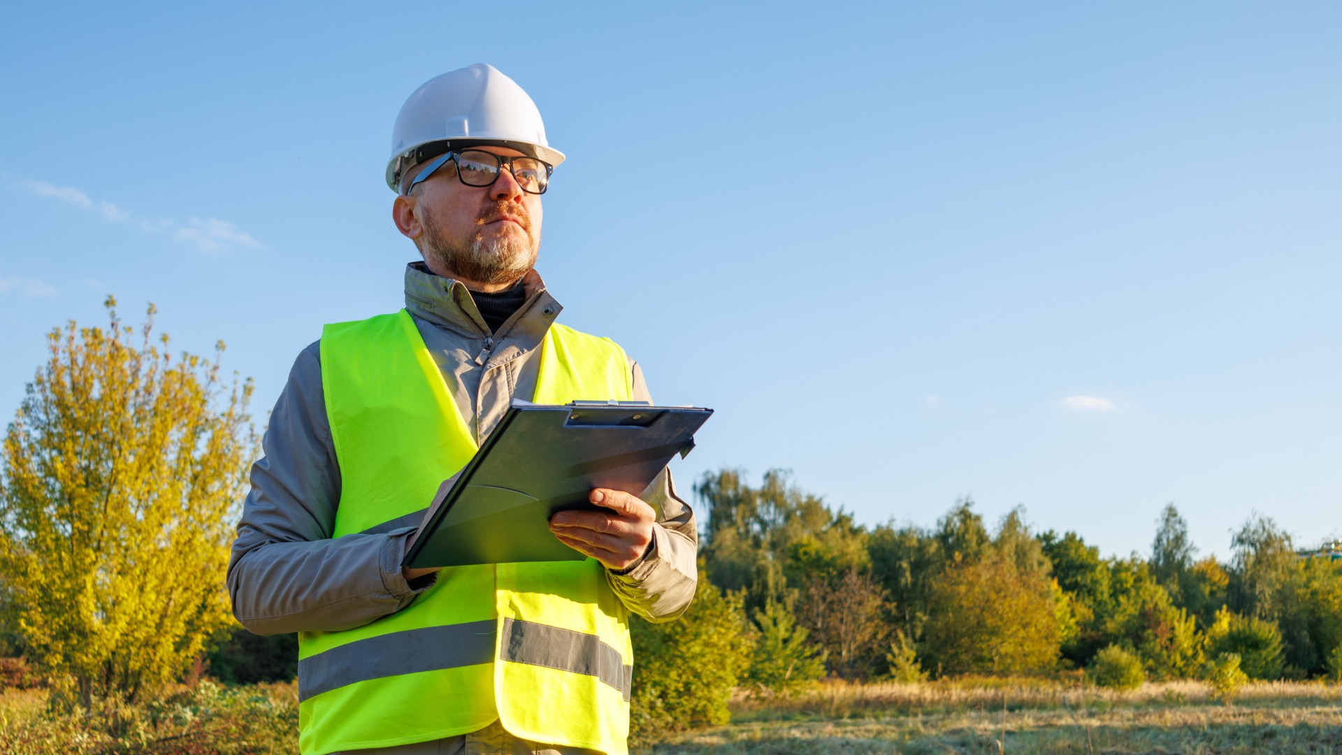 A man wearing a neon-yellow safety vest and a white hard hat holds a clipboard while standing outside.