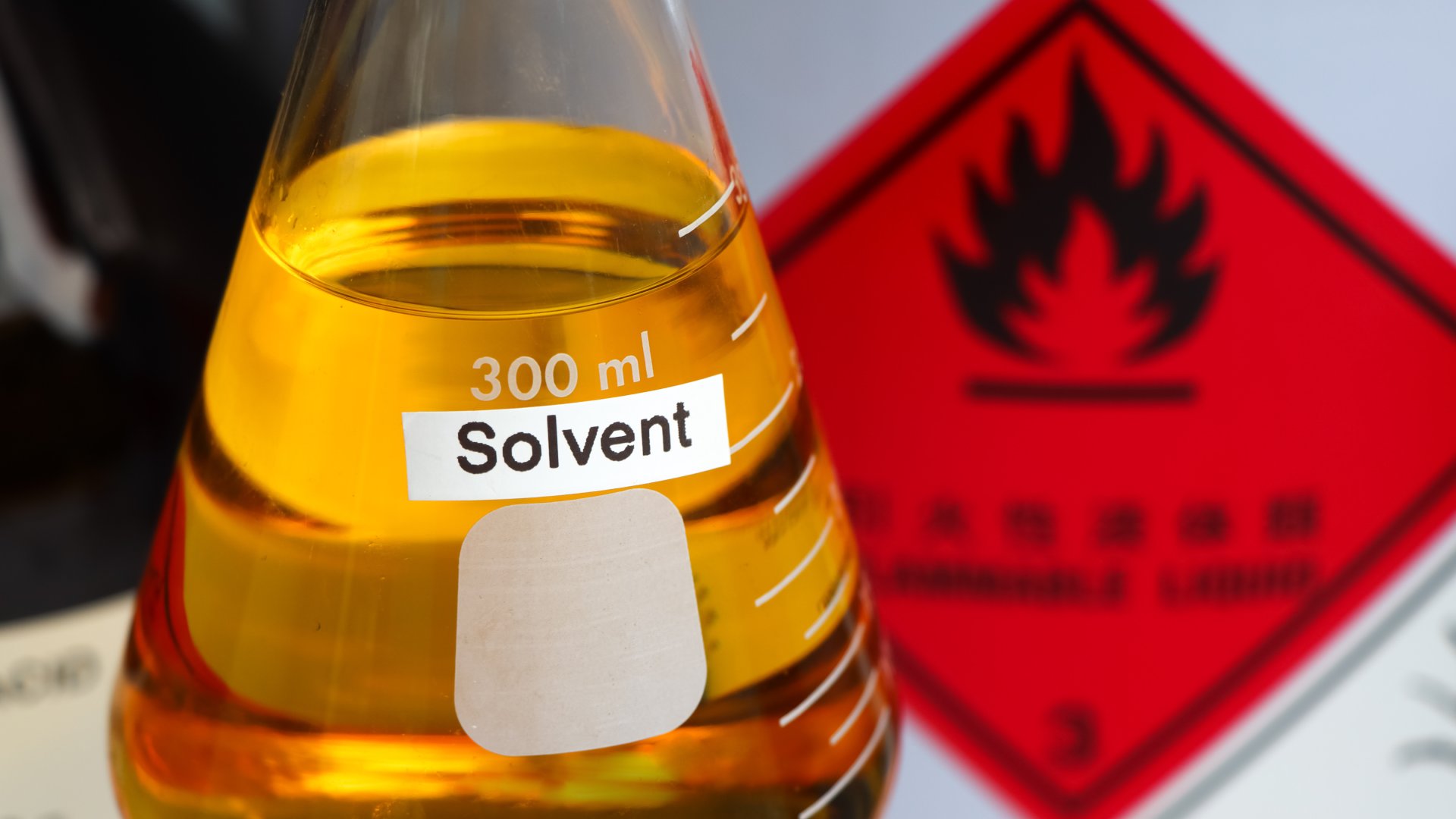 A beaker with a "solvent" label on the front is filled with orange liquid. There is a red flammable sign in the background.