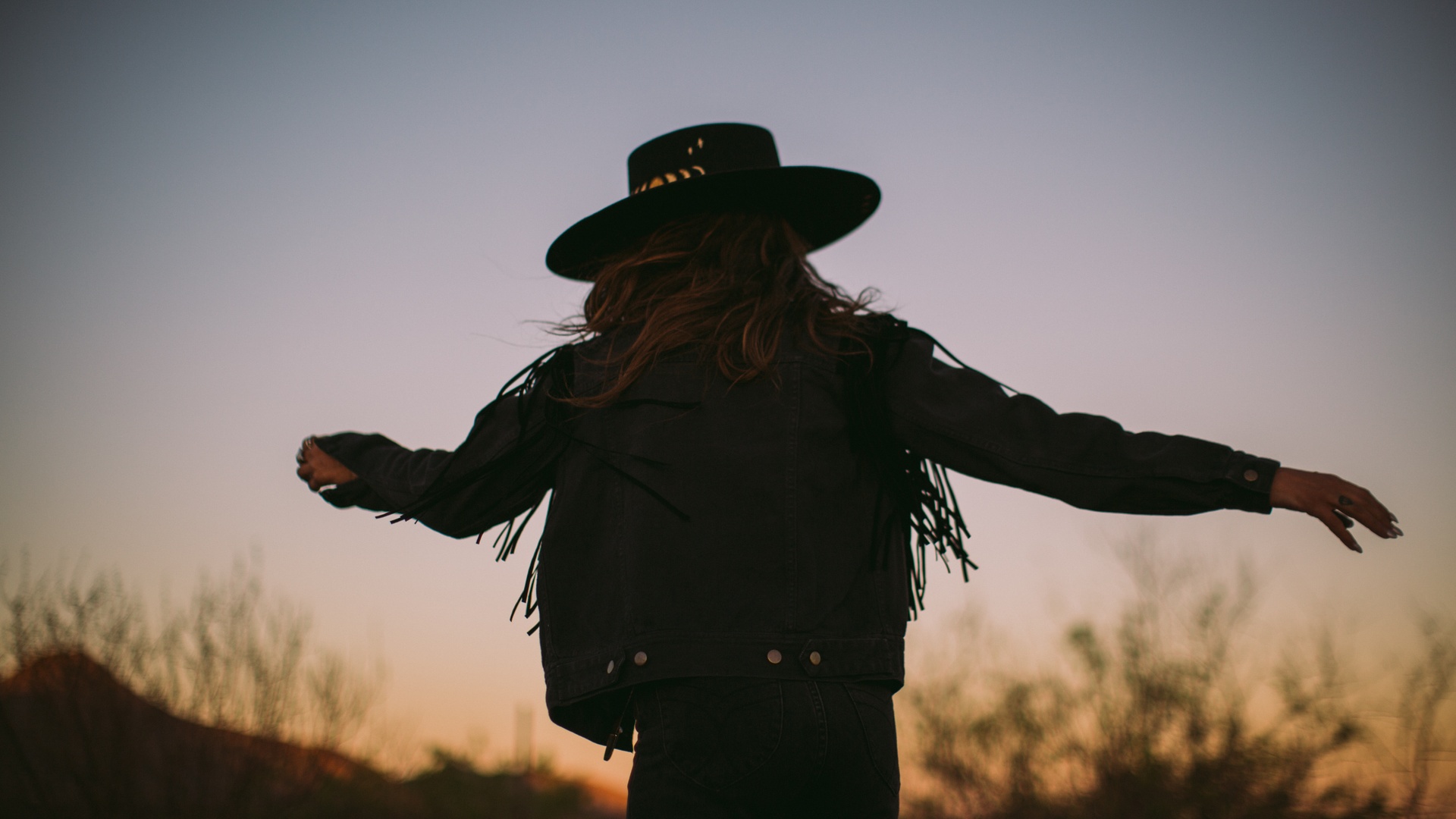 A woman in a western hat and leather fringe jacket is dancing outside, in the sunset, with her back turned towards the camera.