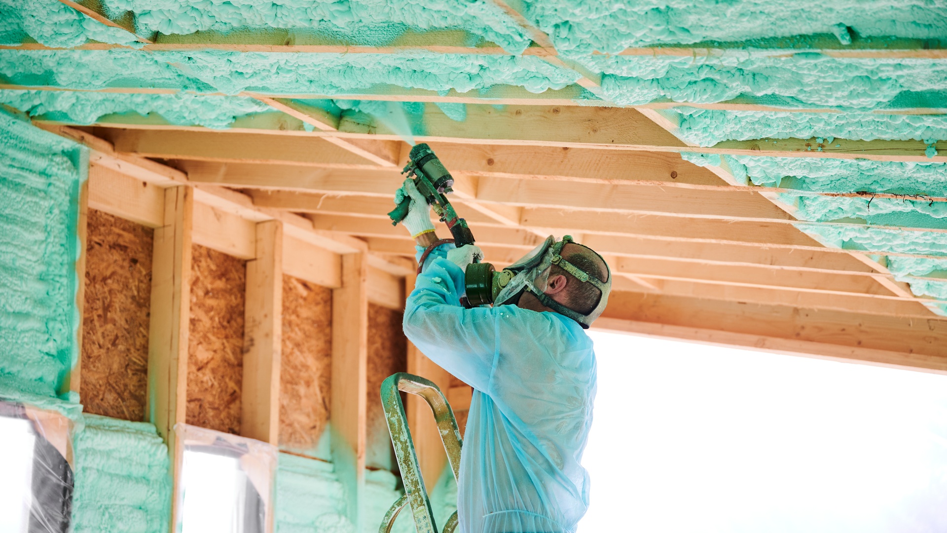A contractor wearing a blue protective suit and mask sprays foam insulation on the wooden frame of a house.