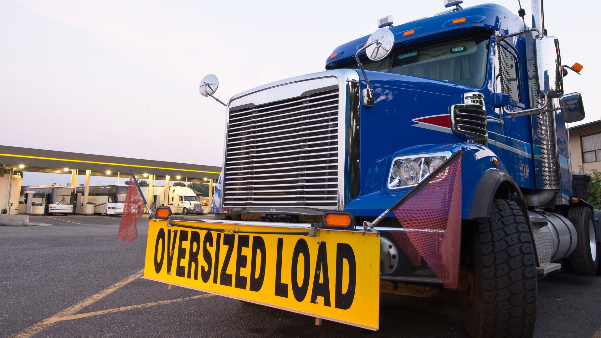 A large truck with a blue cab is parked outside, featuring a yellow sign on the front bumper that reads "Oversized Load."