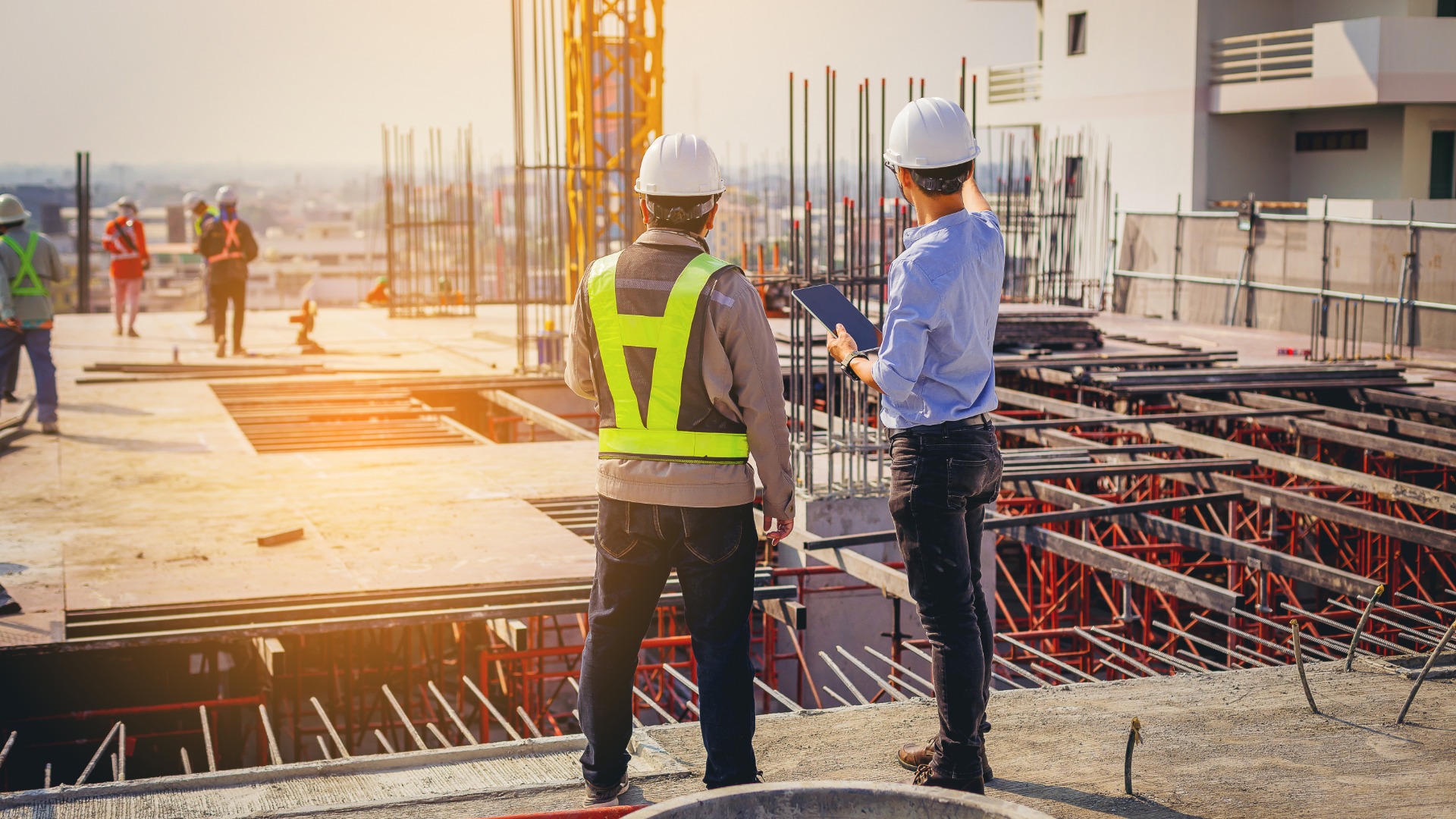 Two people in hard hats standing together while looking at an active construction site. One is holding a tablet and pointing.