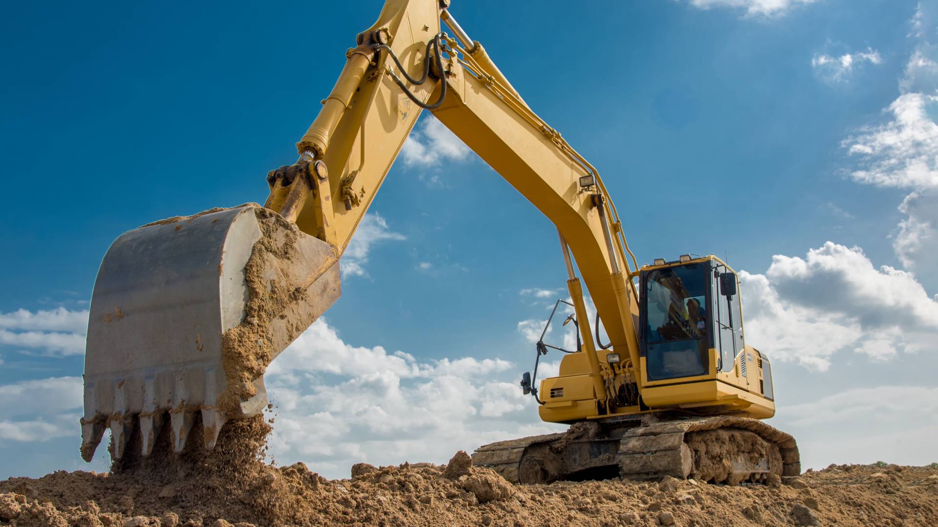 A large, yellow excavator uses a bucket attachment to drop dirt onto more dirt. The blue sky shines behind the excavator.