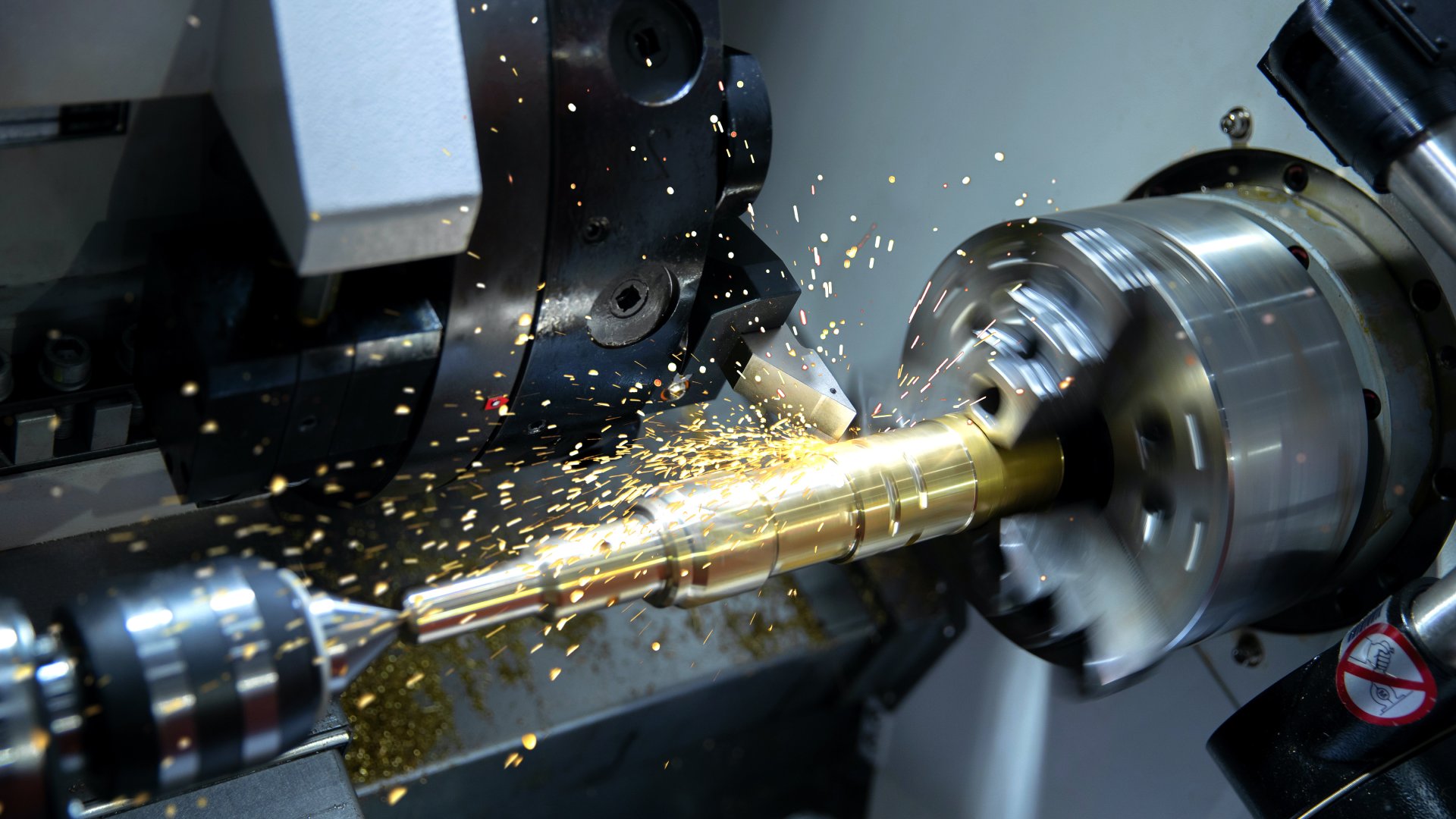 A close-up of a CNC turning machine cutting metal. Sparks fly around the machine as it shapes the material.