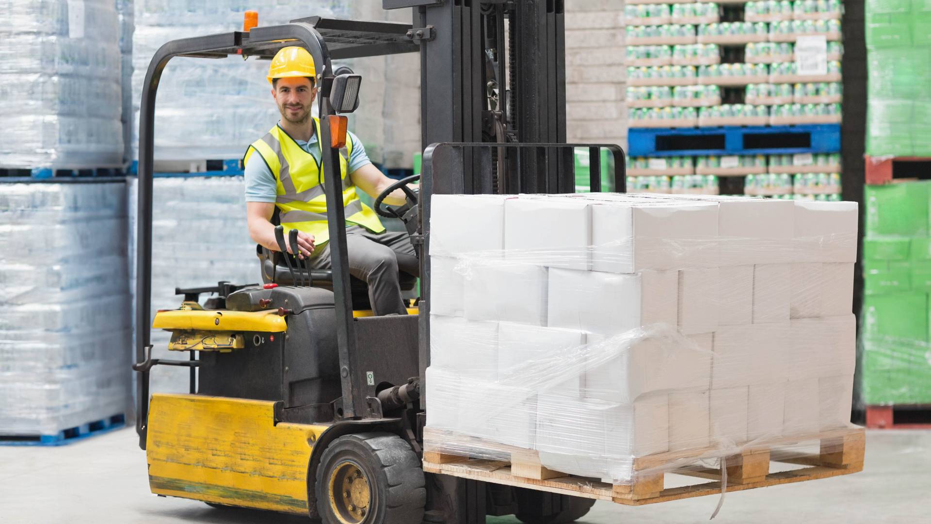  A man wearing a hard hat and safety vest operates a forklift in a warehouse, moving a wooden pallet stacked with white boxes.