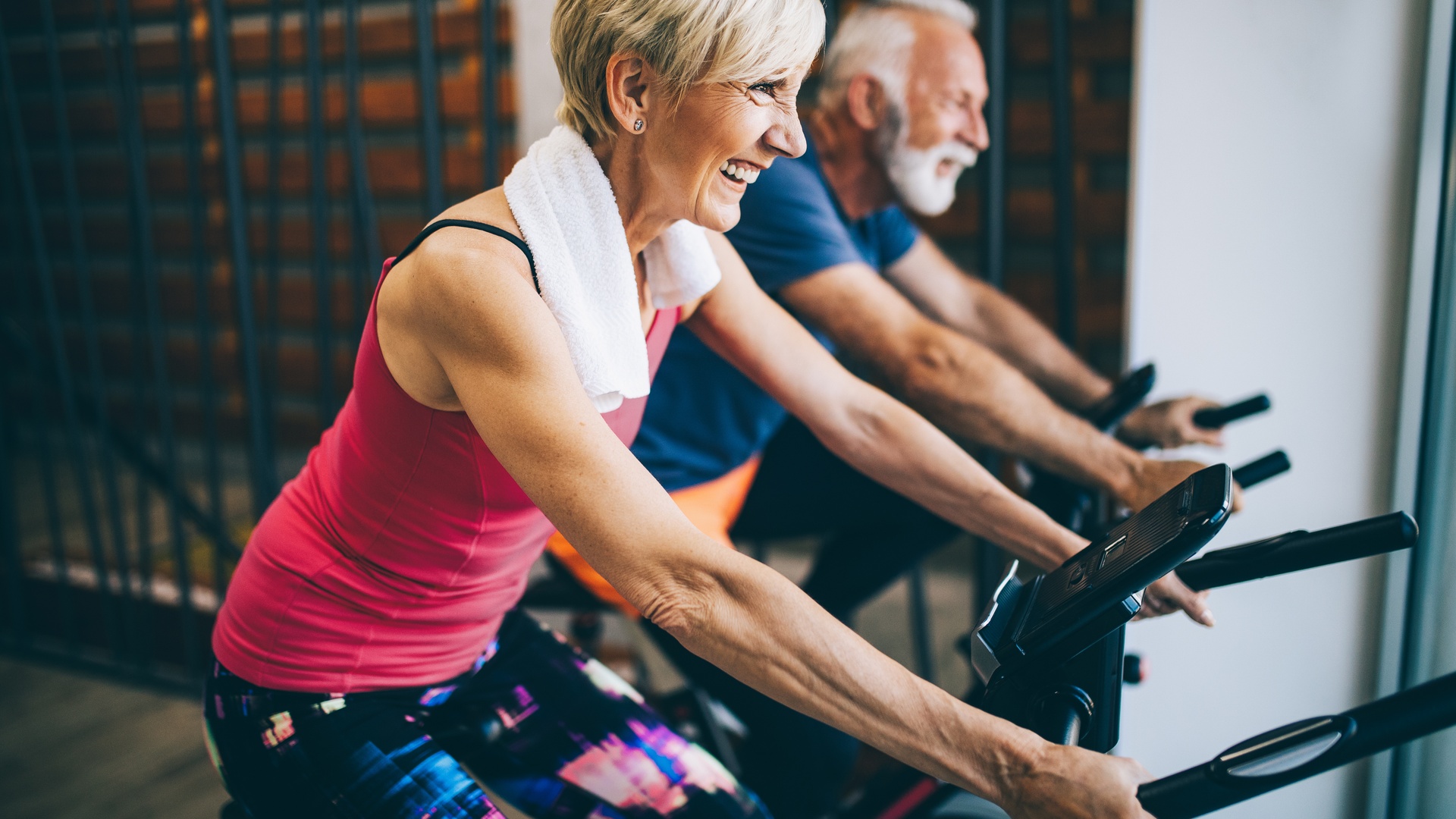 An older man and woman in workout clothes smile as they ride black upright stationary bikes side by side.