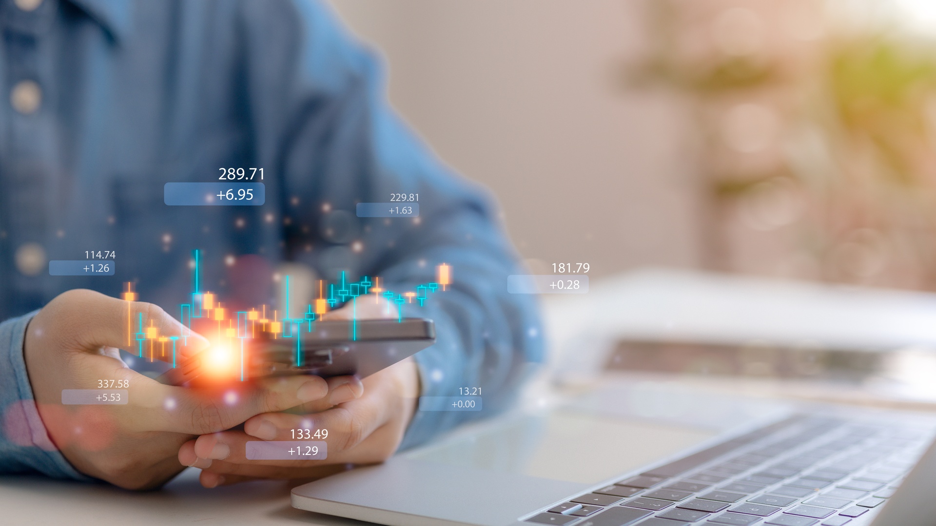 A person holds a smartphone with financial charts overlaid, sitting at a desk with a laptop in the background.