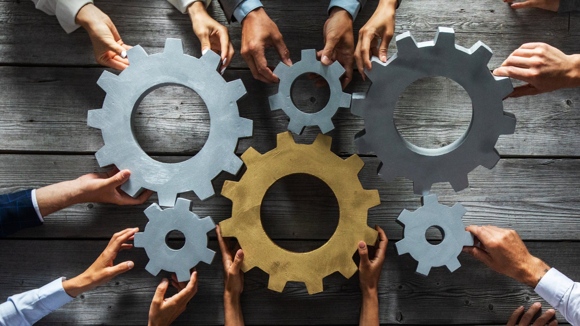 A close-up of a group of businesspeople's hands joining to turn silver and gold metal industrial gears.