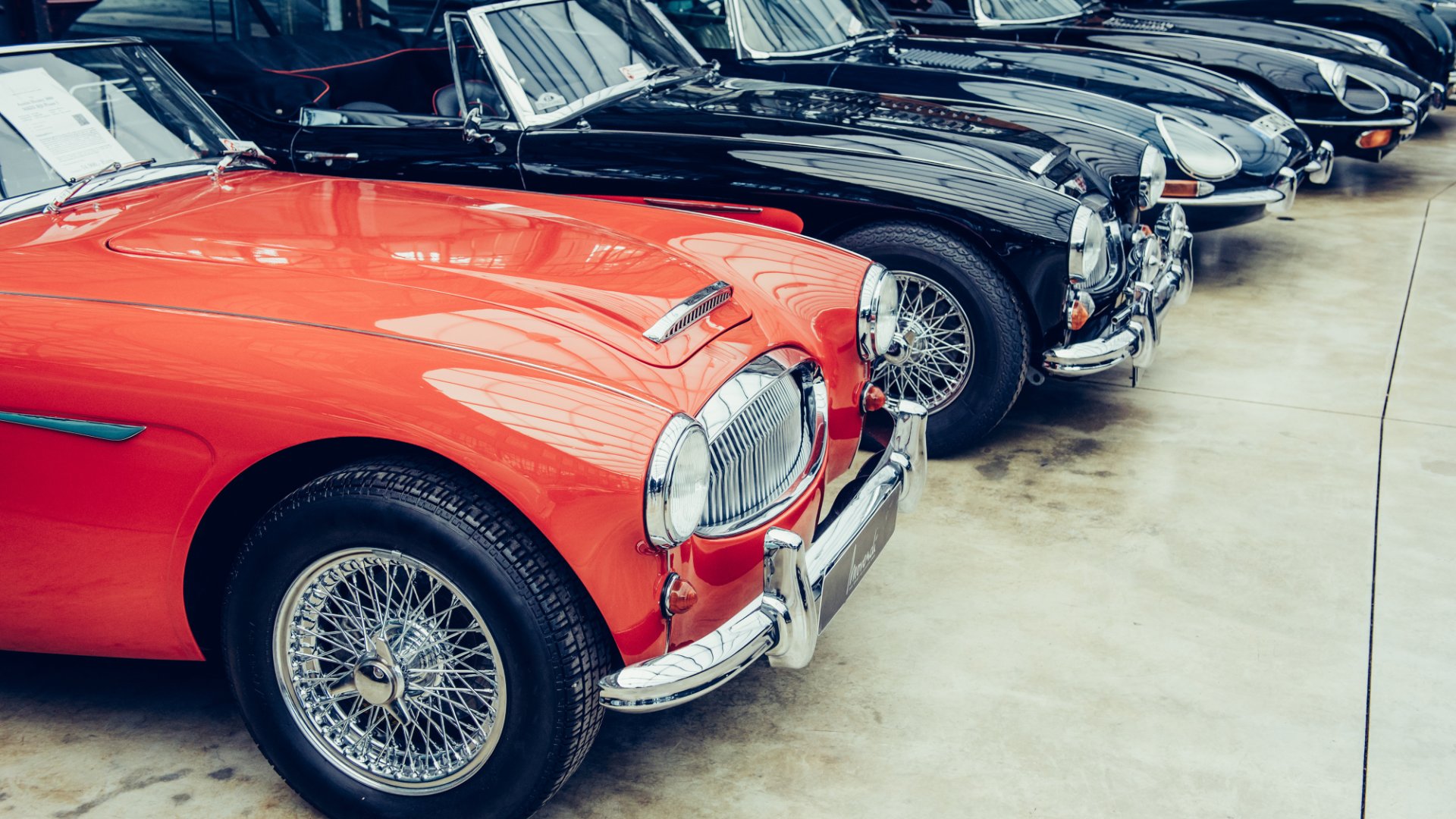 Row of classic convertibles in a showroom, led by a red car with chrome bumper and wire-spoke wheels.