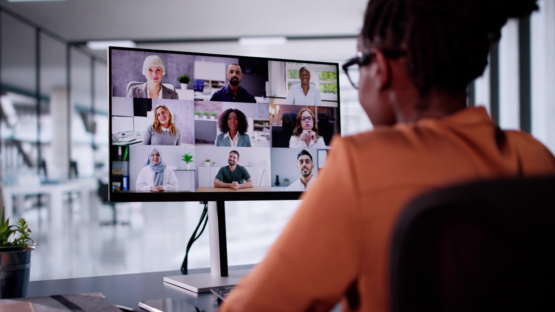 An over-the-shoulder view of a woman in an orange blouse in an office having a virtual meeting with coworkers.