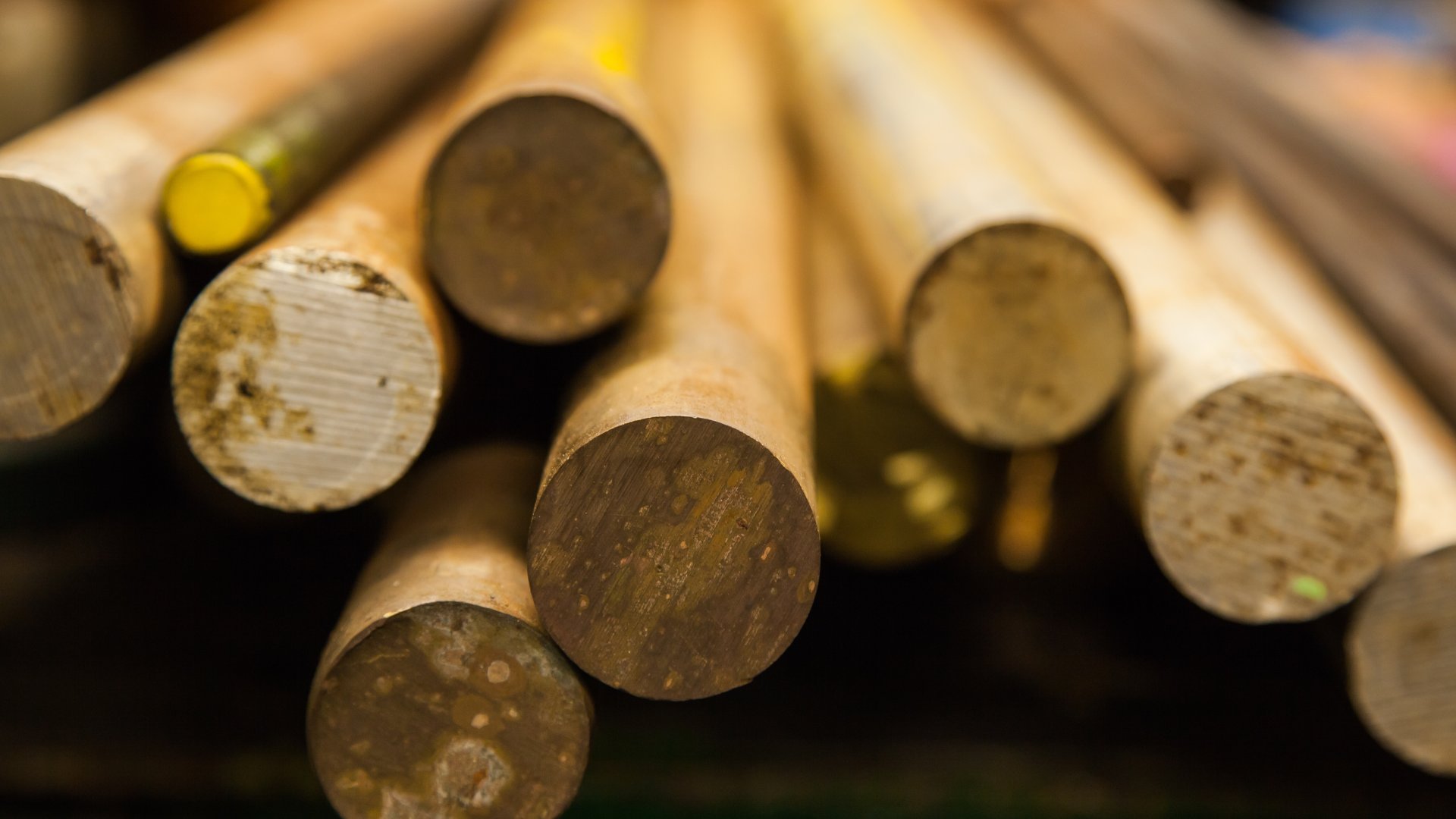 A close-up view of several large bronze round threaded bars, stacked on a green shelf under soft lighting.