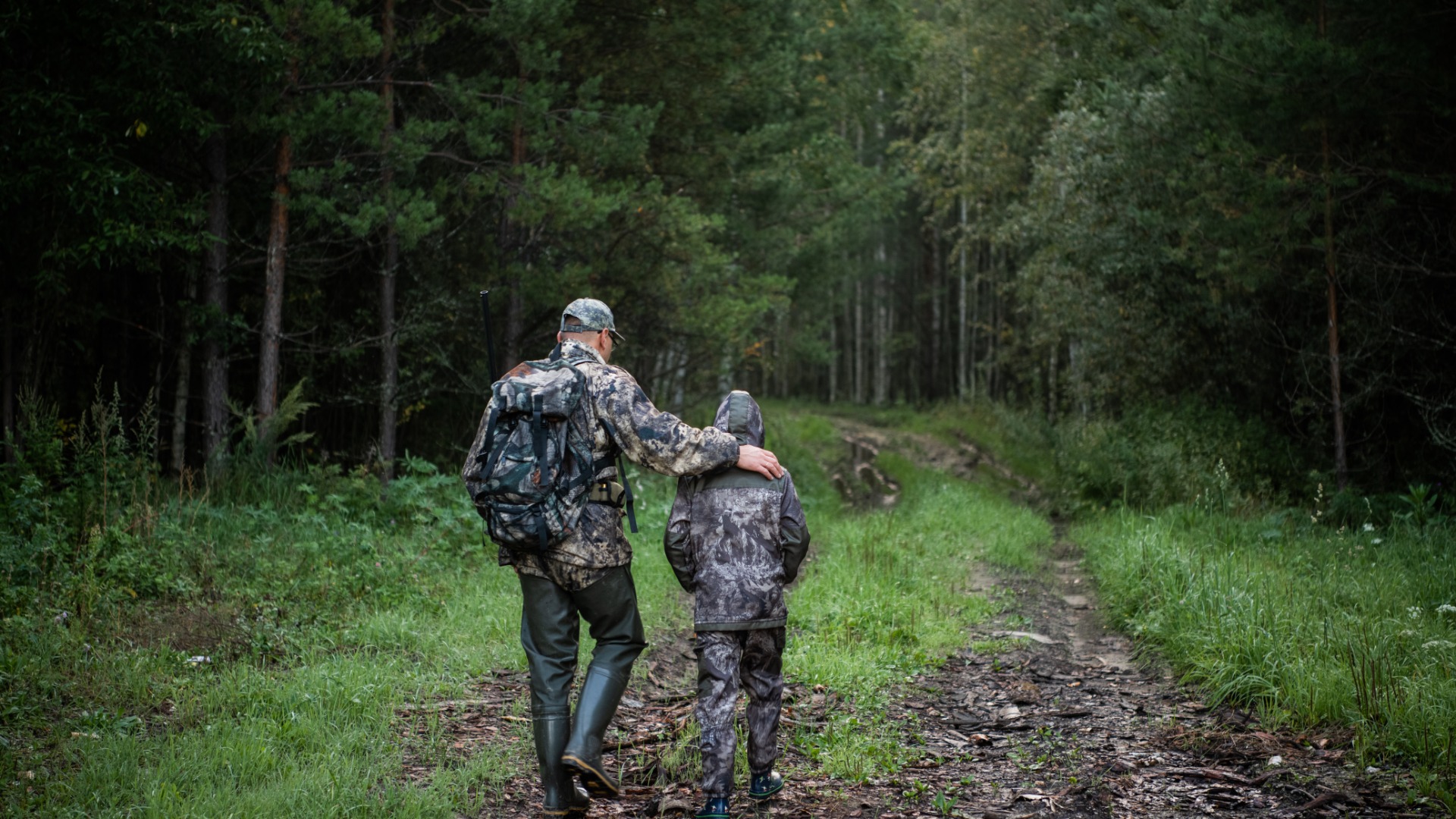 A man and his son are walking through a wooded area. They are both wearing camouflage clothing.