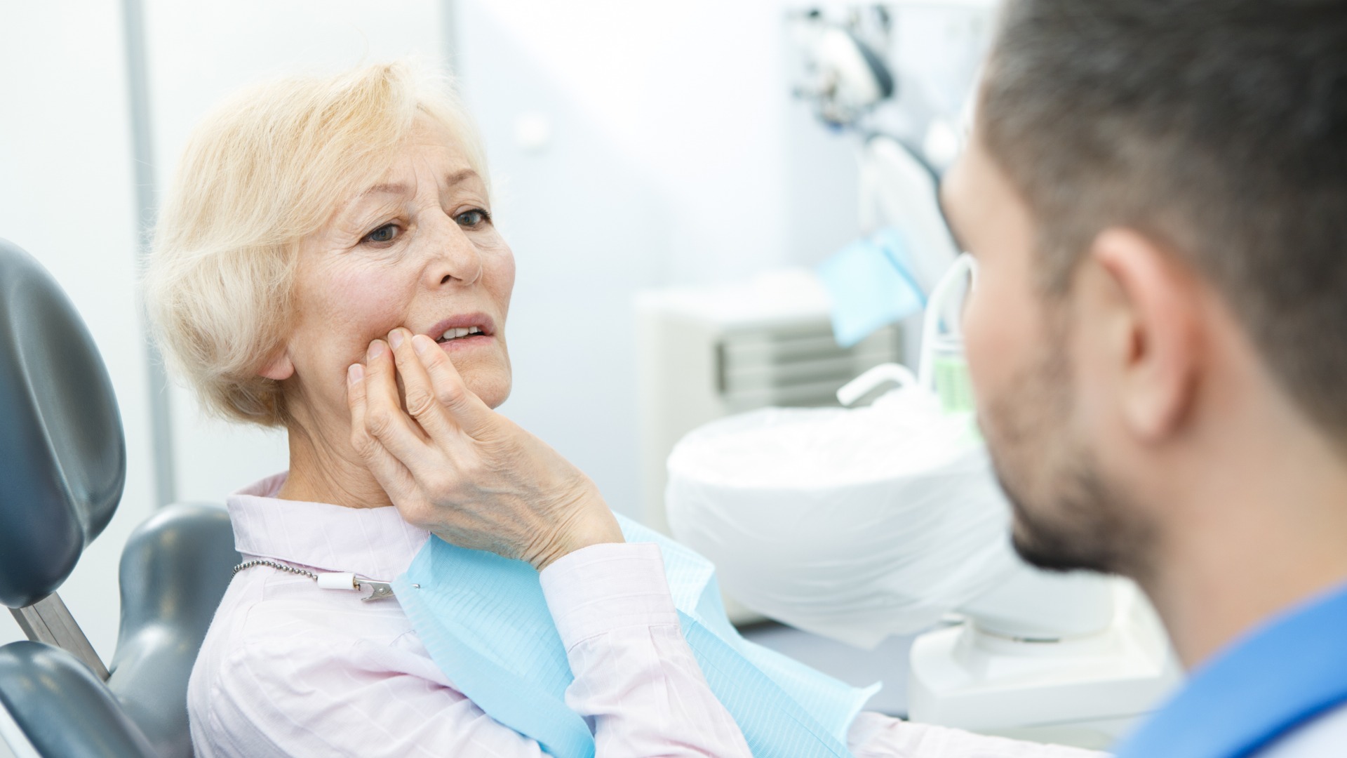 A senior woman in a dentist's chair, looking at her doctor. She holds a hand to her jaw with a pained expression.