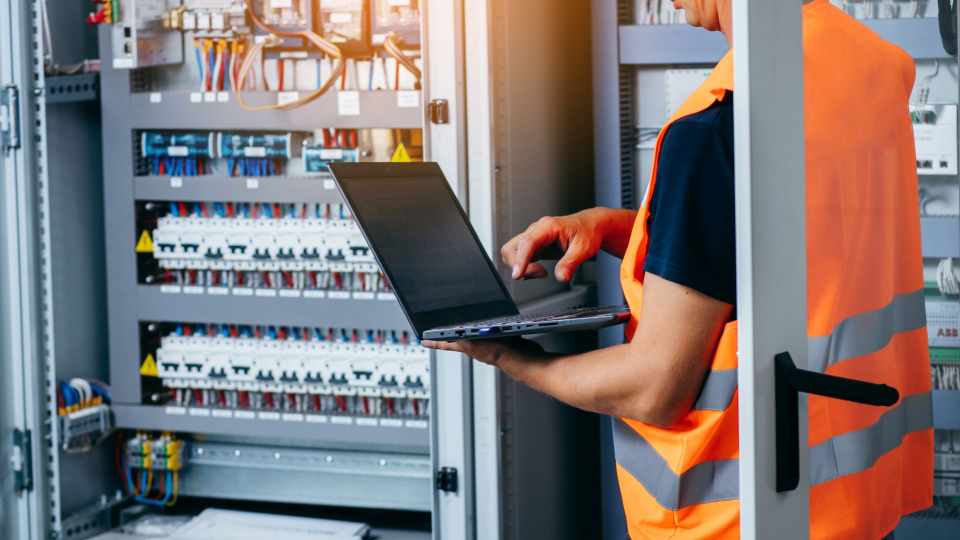 An electrician is repairing an electricity power station and checking the control cabinet with a laptop in his hands.