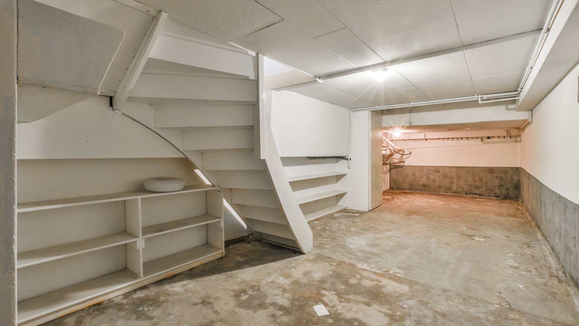 A home's partially finished basement has the original cement flooring with a finished ceiling and walls with some shelves.