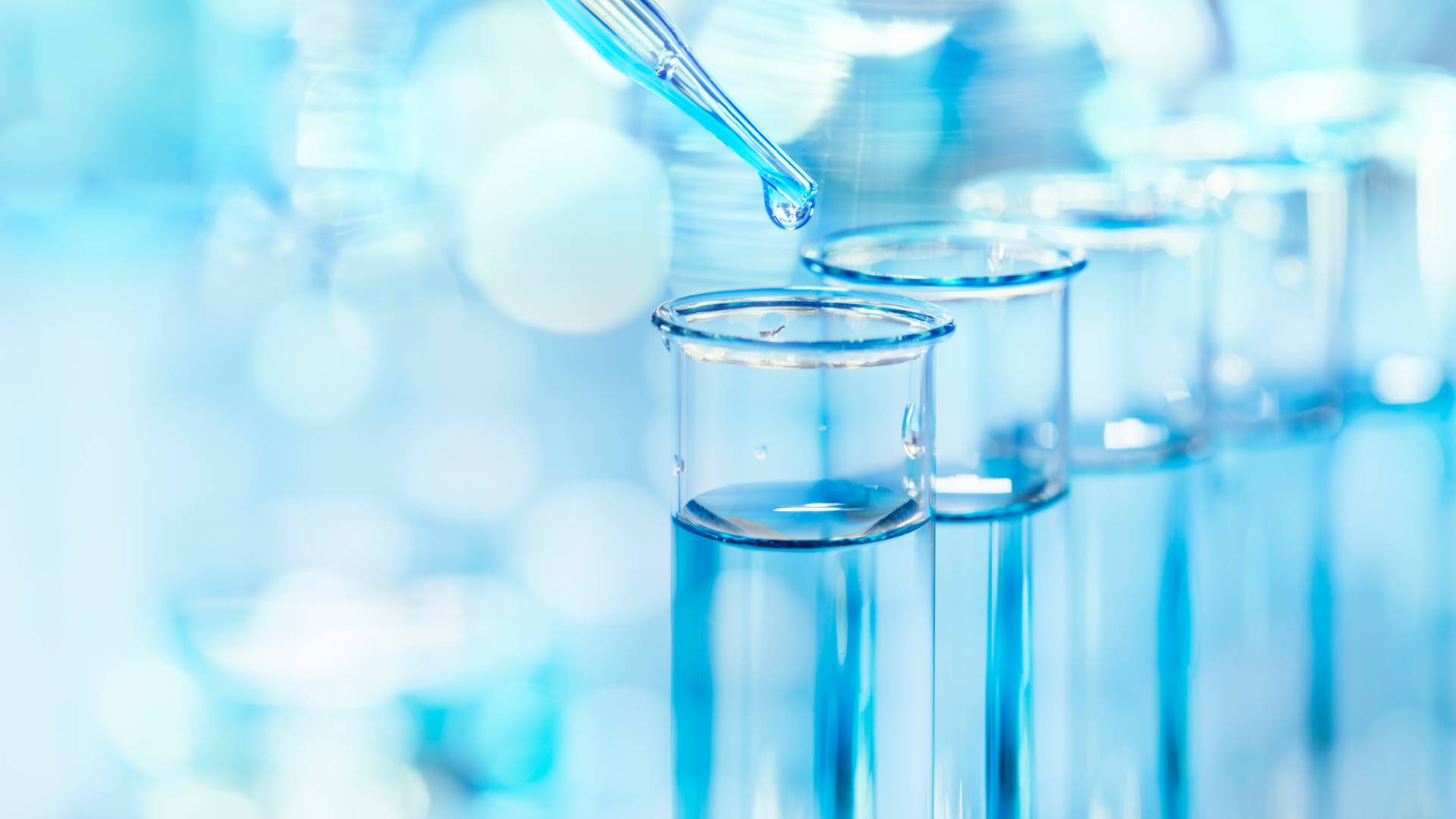 A transfer pipette filling a test tube. A row of several blue-liquid filled test tubes are in line behind the first tube.