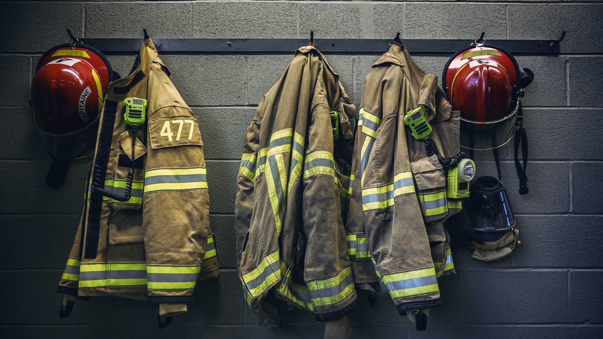 Three firefighter jackets hang alongside two red helmets on black hooks attached to a black cement wall.