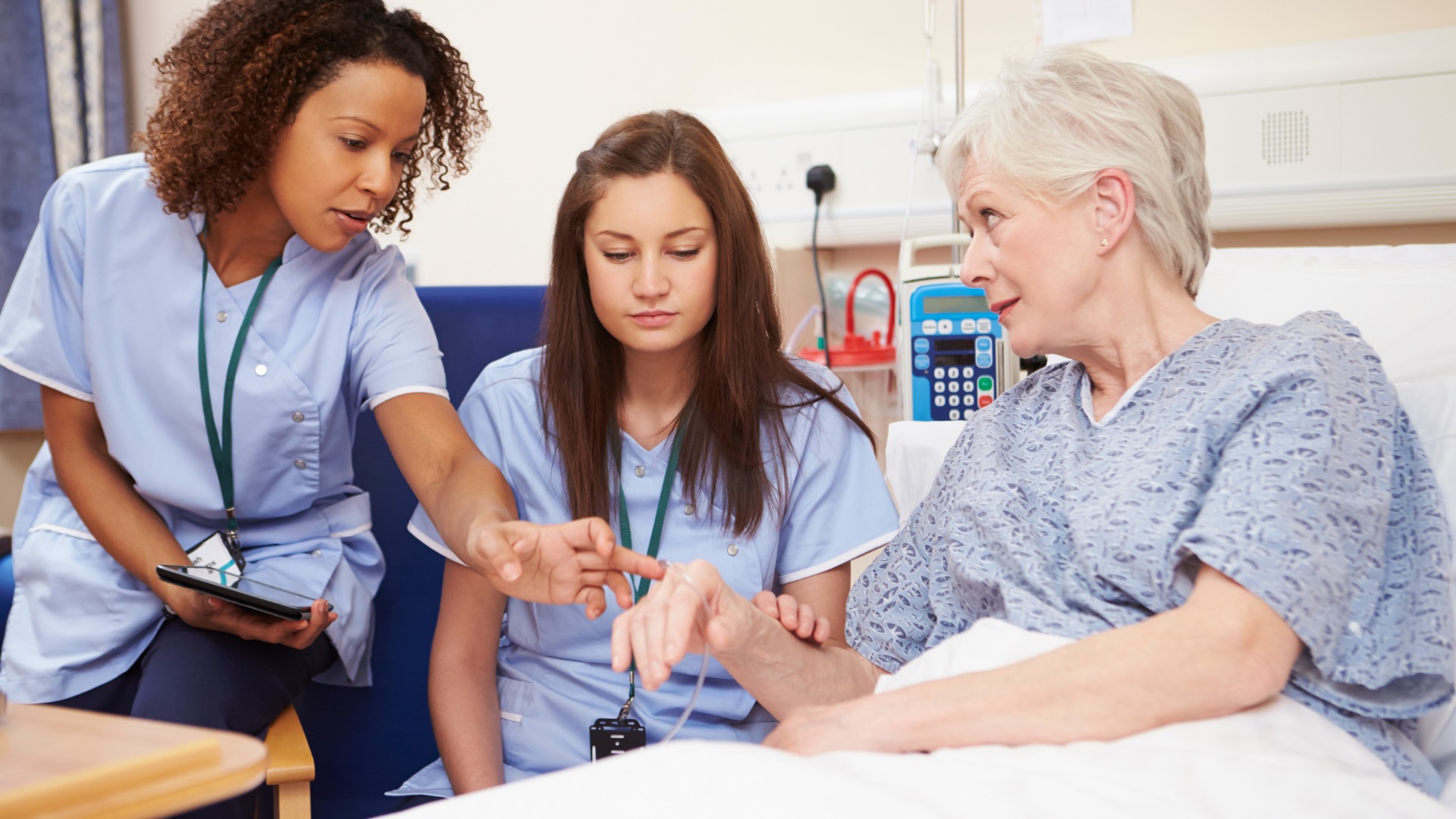 A senior nurse instructs a student nurse at a patient's bedside inside a hospital room during a clinical training visit.