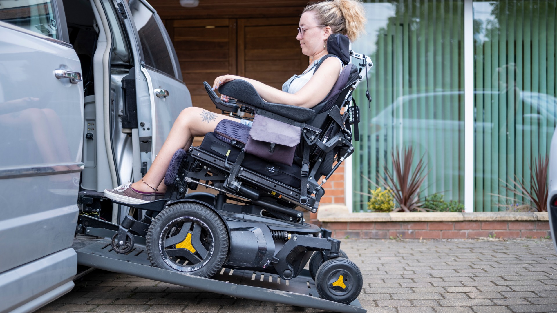 A woman in an electric wheelchair uses a ramp to get into an accessible van. The van is parked on a brick driveway.