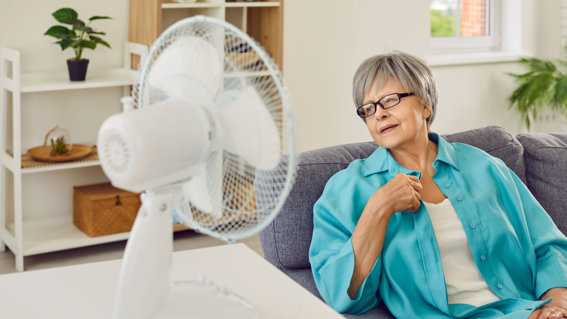 A woman in a blue shirt wearing glasses sitting on a gray sofa beside a white electric fan in a bright living room.