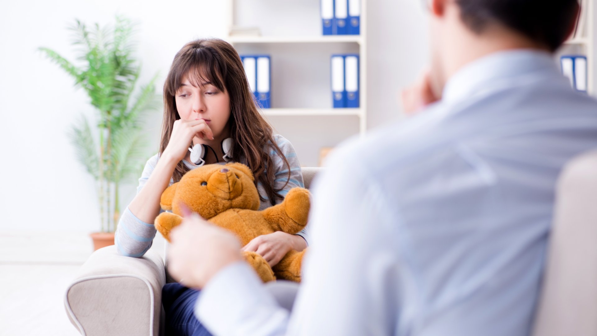 A young girl sitting with a teddy bear in front of a talk therapist. She has her hand on her mouth and she's looking down.