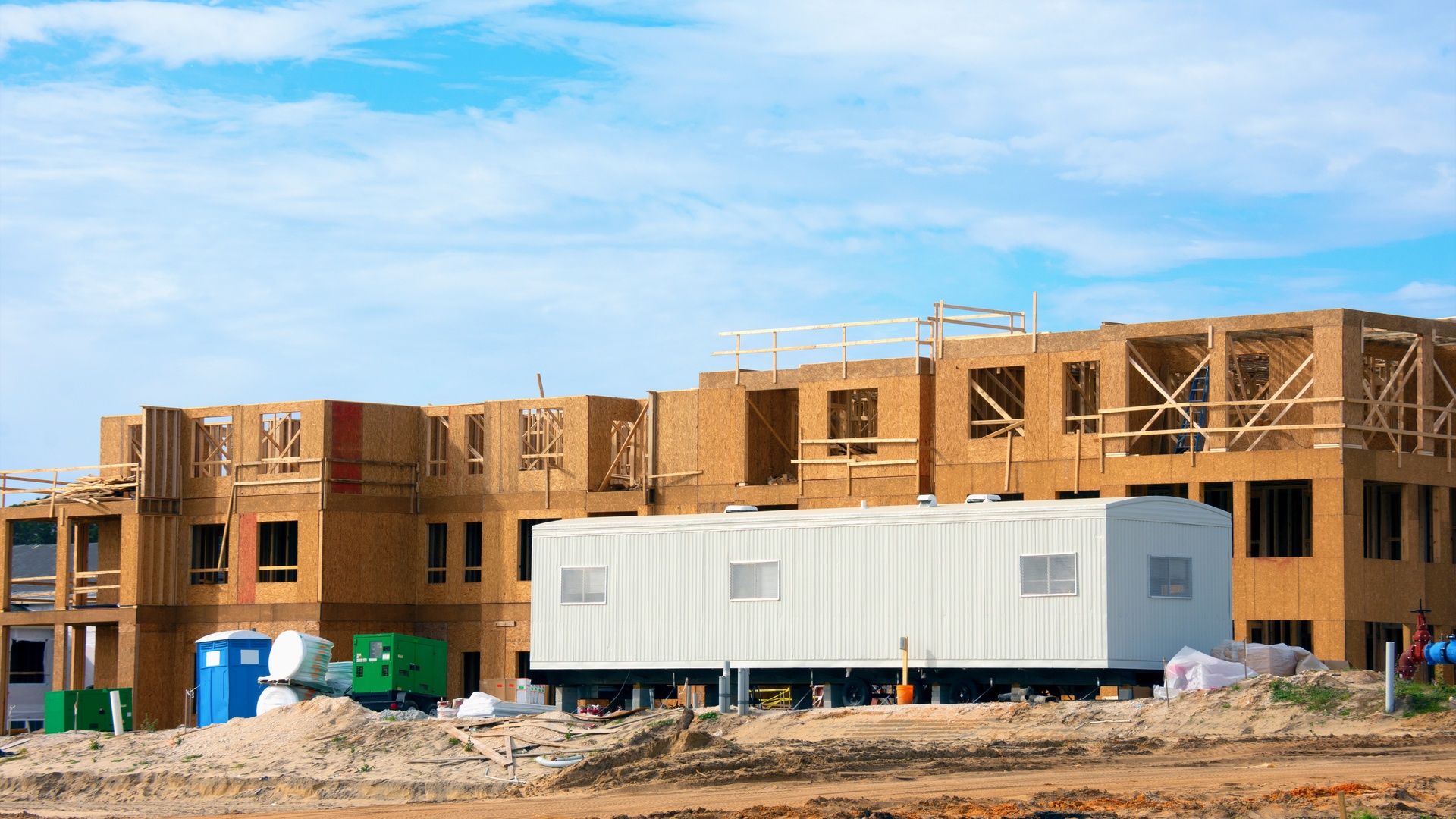 A remote construction job site with a white mobile trailer unit in the front of the build. Dirt surrounds the building.