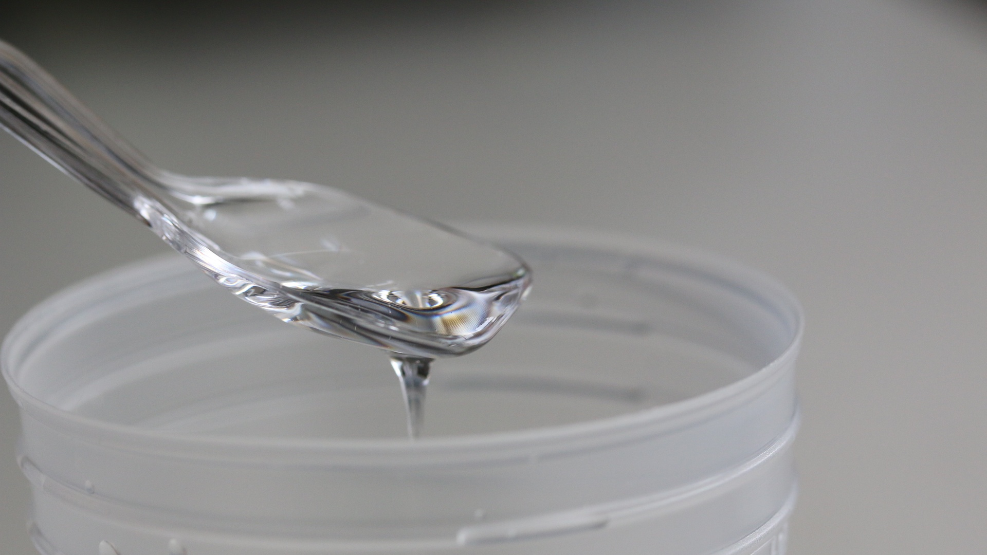 Clear resin drips from a transparent spatula into a cylindrical container, set against a blurred gray background.