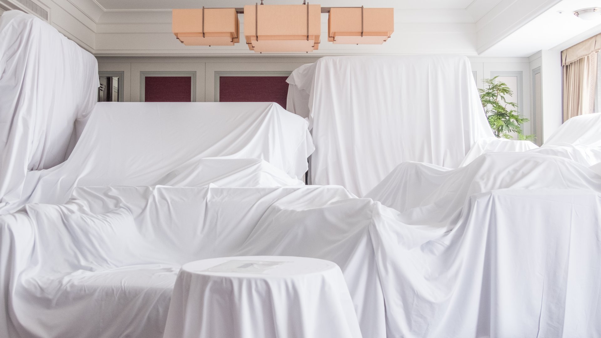 White sheets cover the sofas, chairs, and tables in a bright room with tall windows and a beige chandeliers.