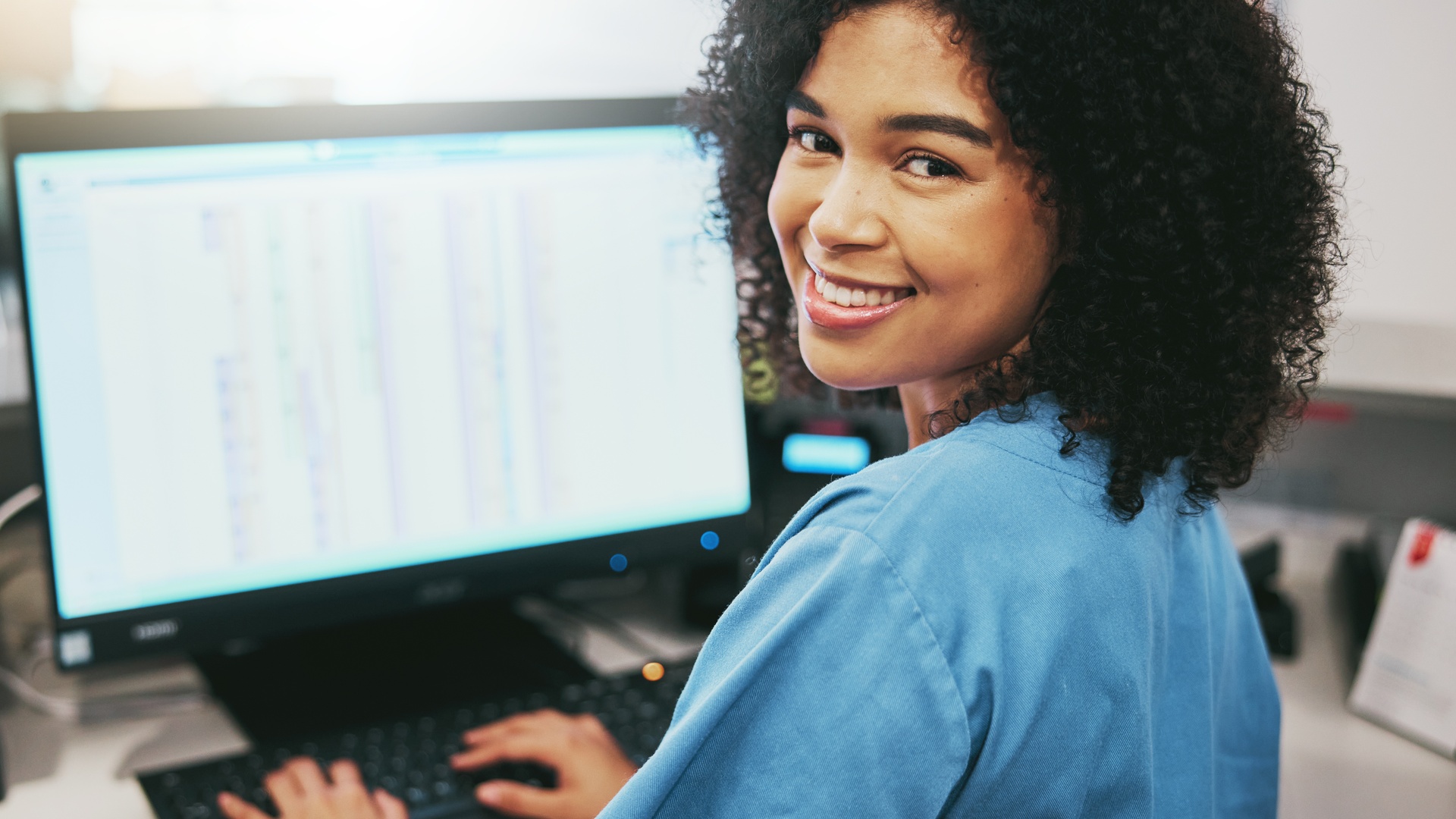 A medical office staff member smiles at her desk as she works on a computer, symbolizing efficient healthcare administration.