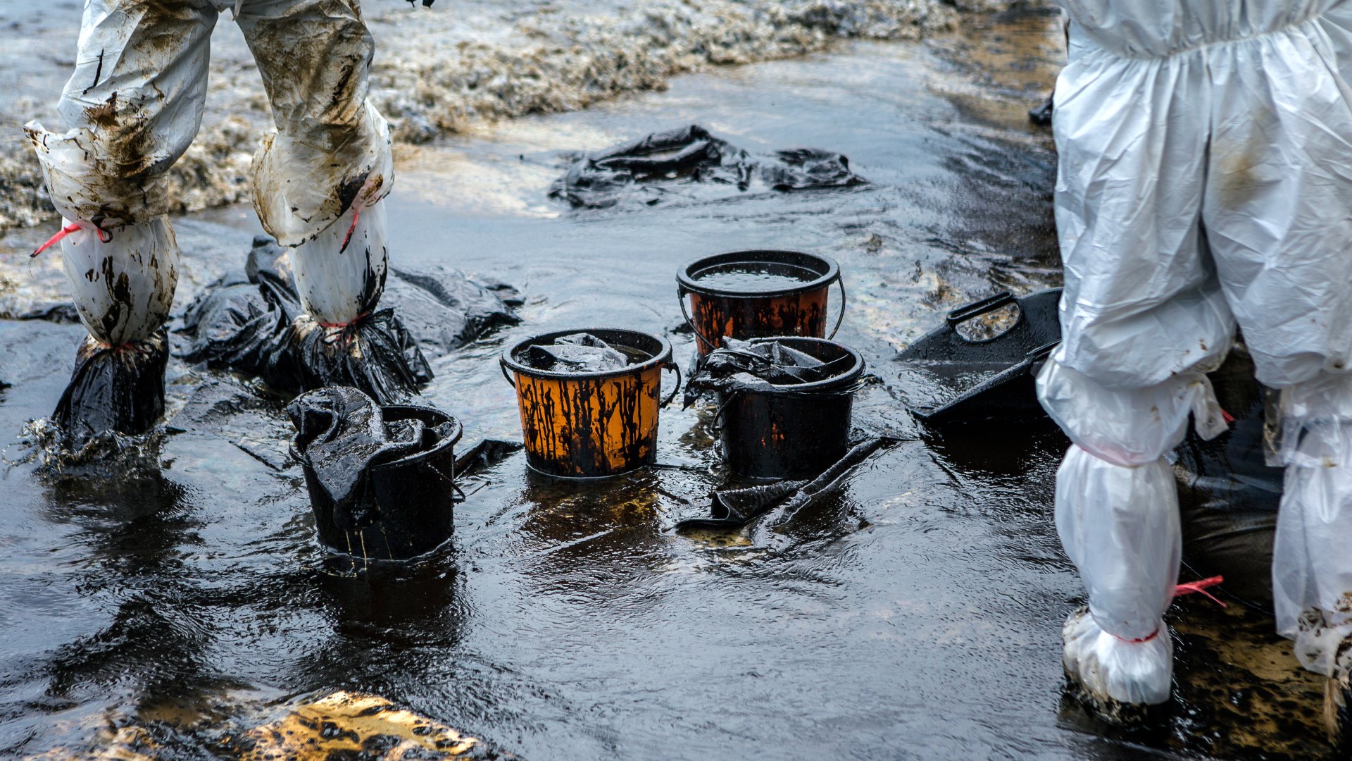 Two crew members wade an oil spill in protective gear as they use four buckets to begin recovery cleanup.
