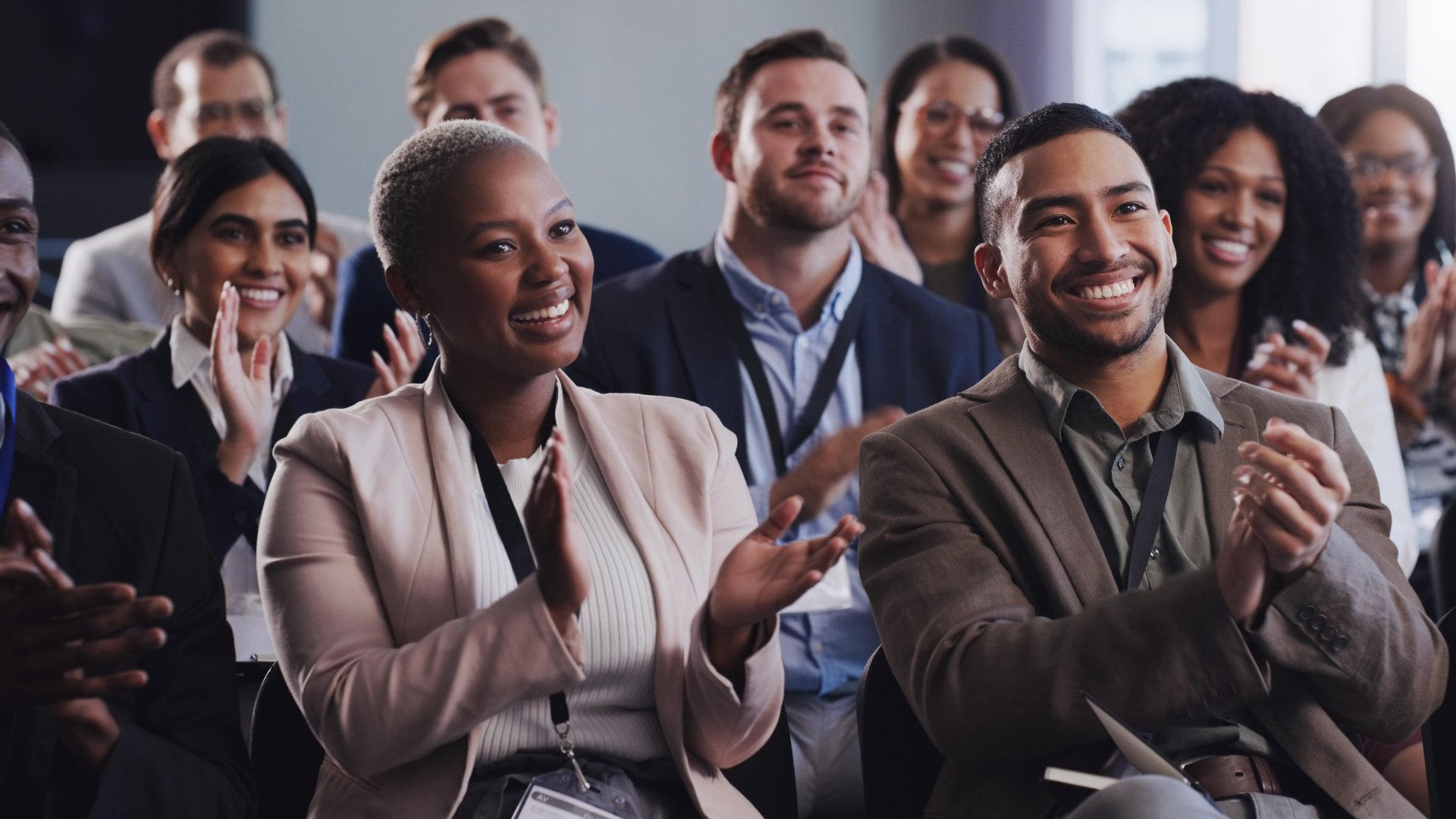 A group of smiling people in business attire sitting in rows and clapping in a bright indoor conference room.