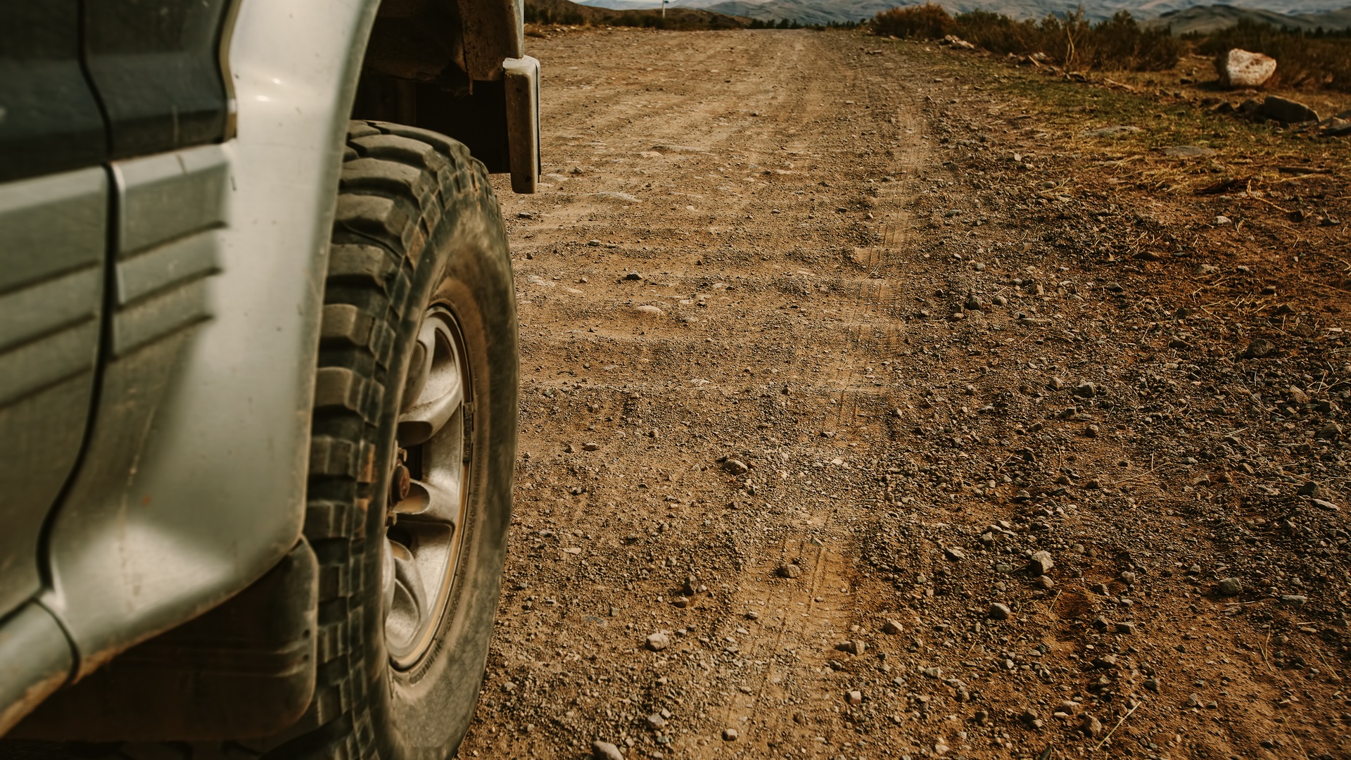 A close up of a car's tires as it drives across a brown dirt road. In the far distance there are shrubs and hills.