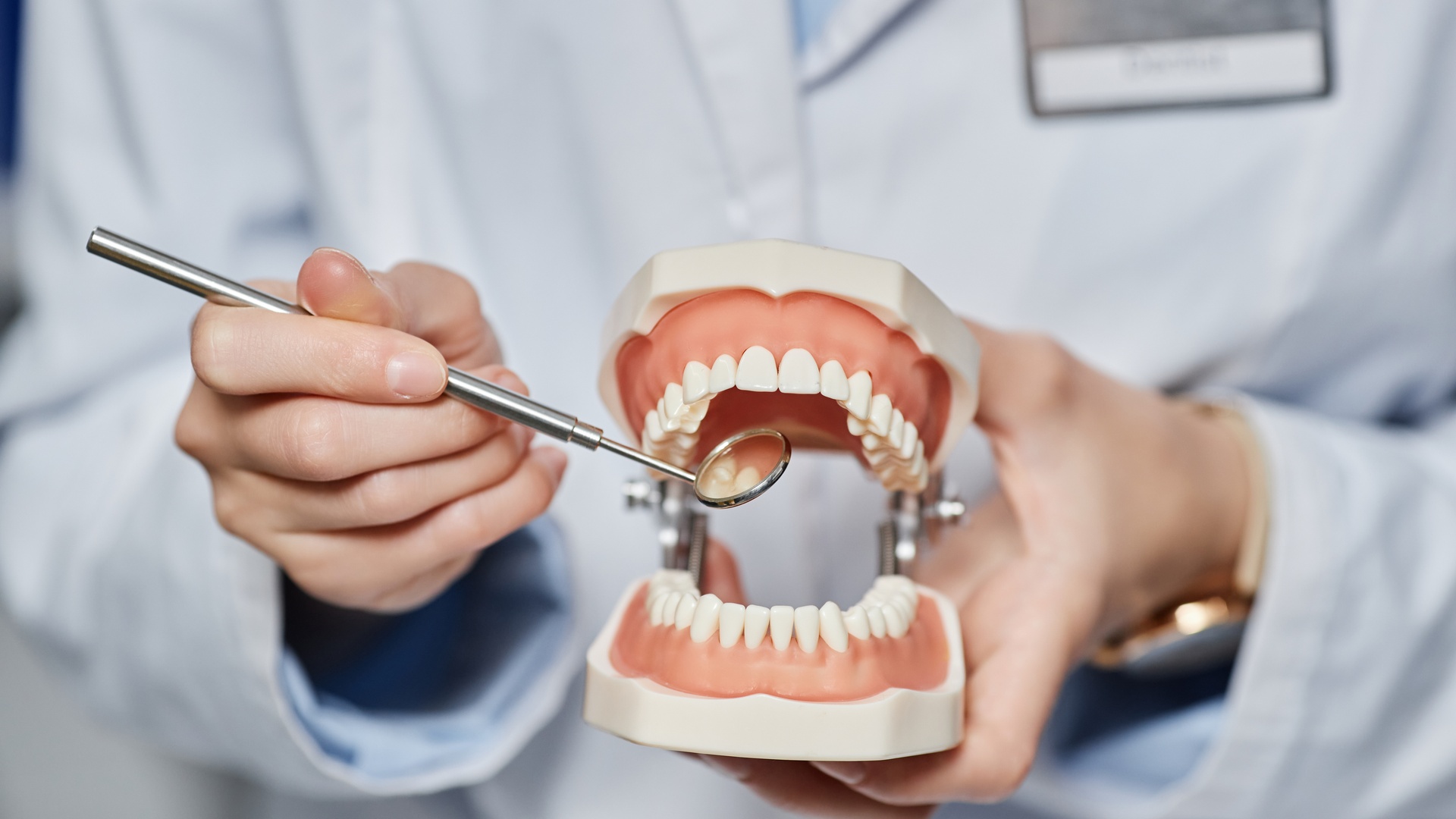 A person wearing a white coat and nametag holding a magnifying glass up to a tooth model in their hand.