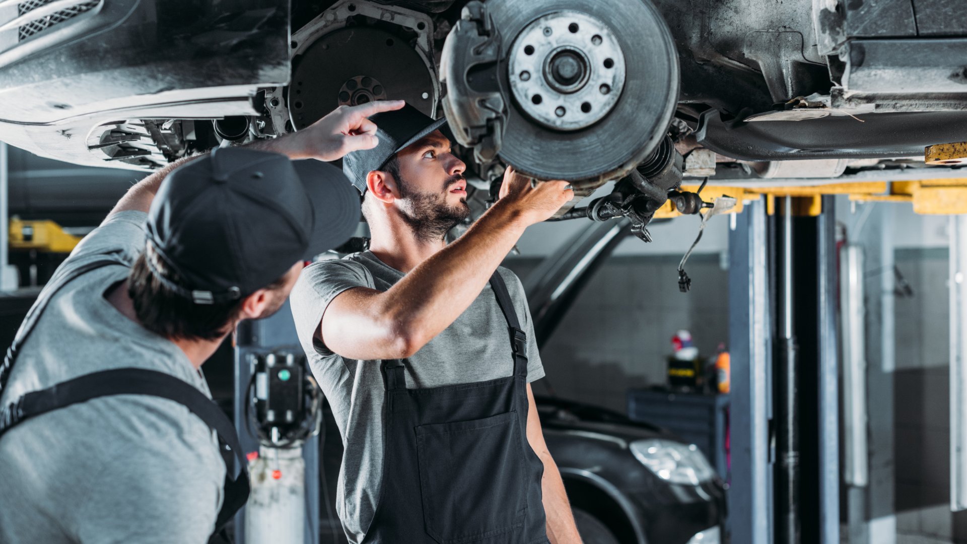 Two mechanics working on the underside of a car in an auto repair shop. The car is raised above the mechanics.