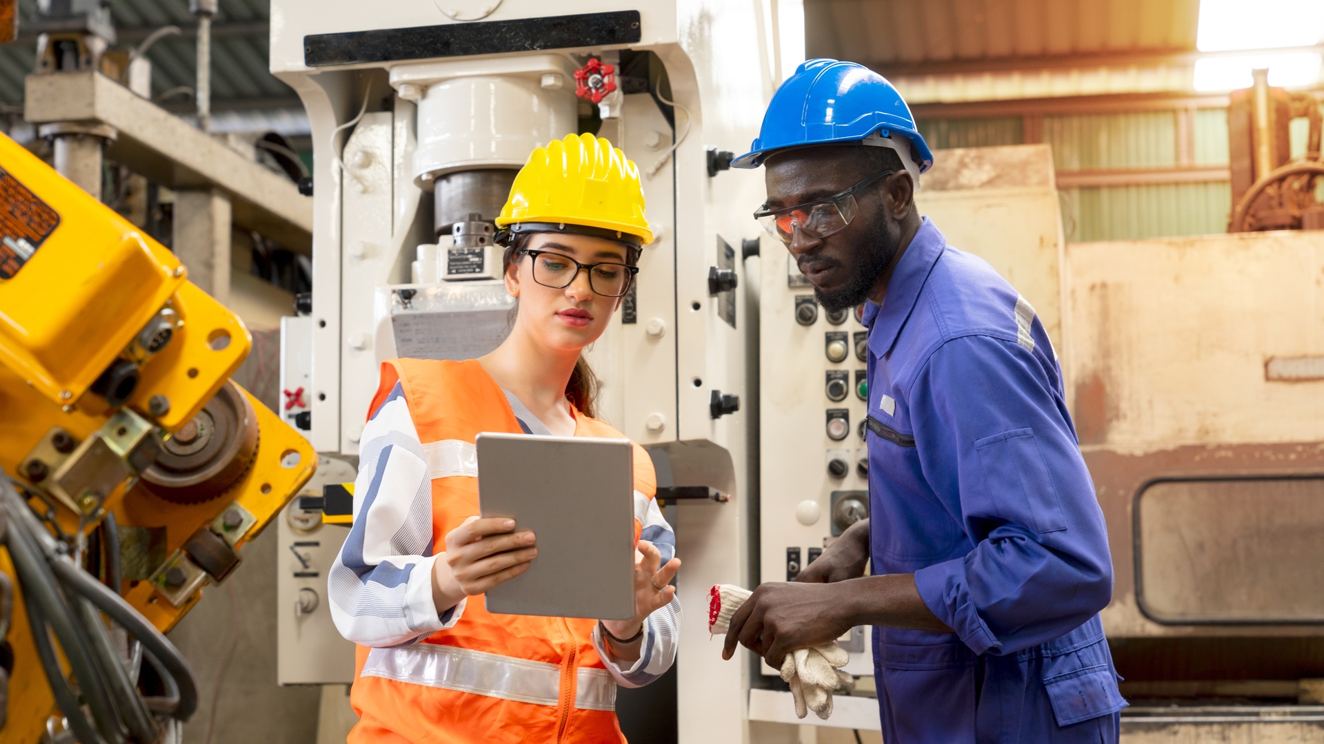 Two employees wearing hard hats in front of a machine in a manufacturing facility. One employee holds a tablet.