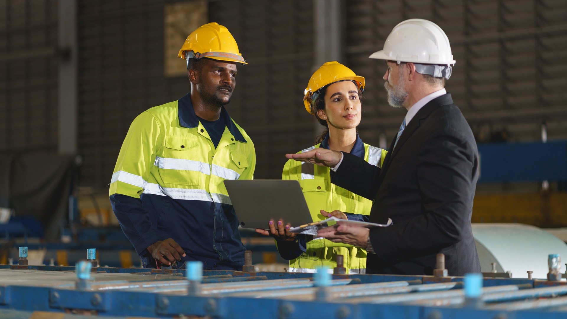 A man in a business suit and hard hat talking to two factory workers. Both of them are in hard hats and reflective attire.