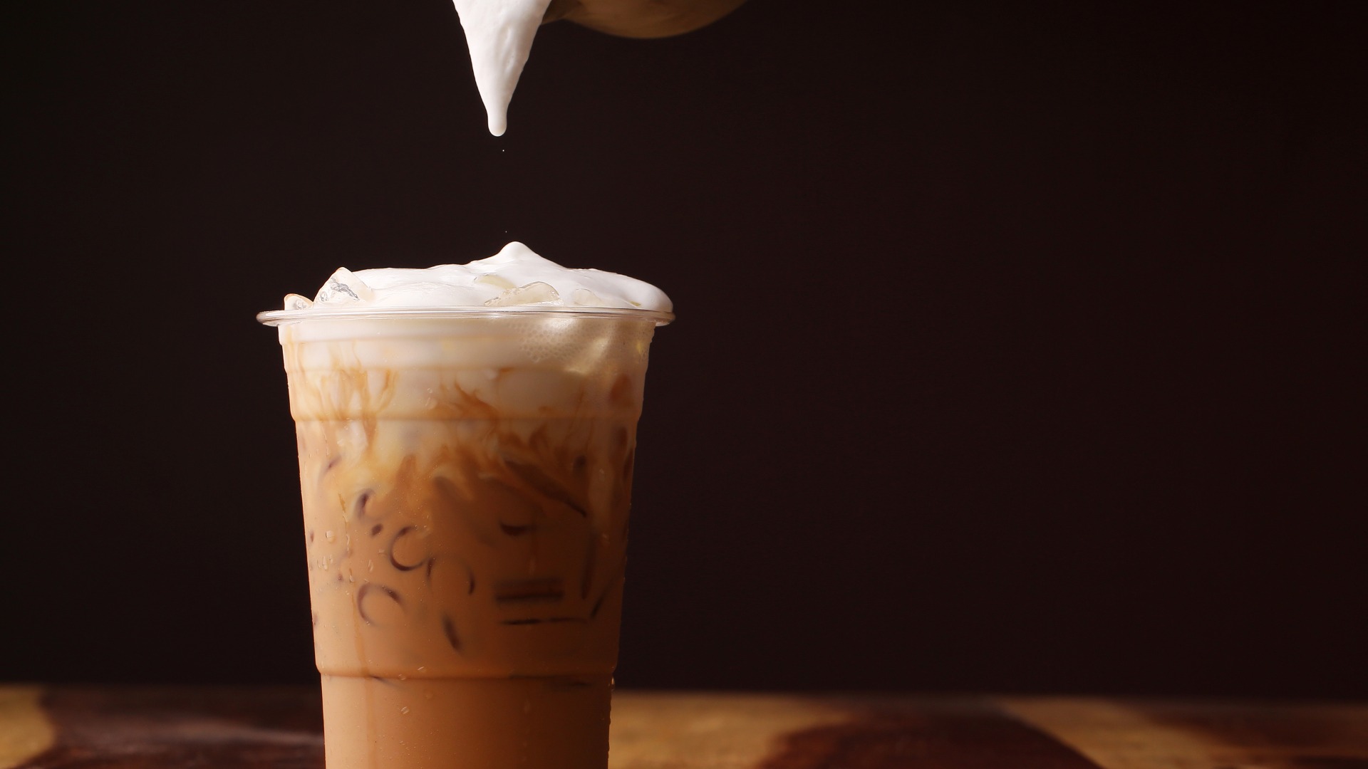 An iced coffee in a plastic to-go cup sits on a table with a pitcher over the top dropping the cold foam on top.