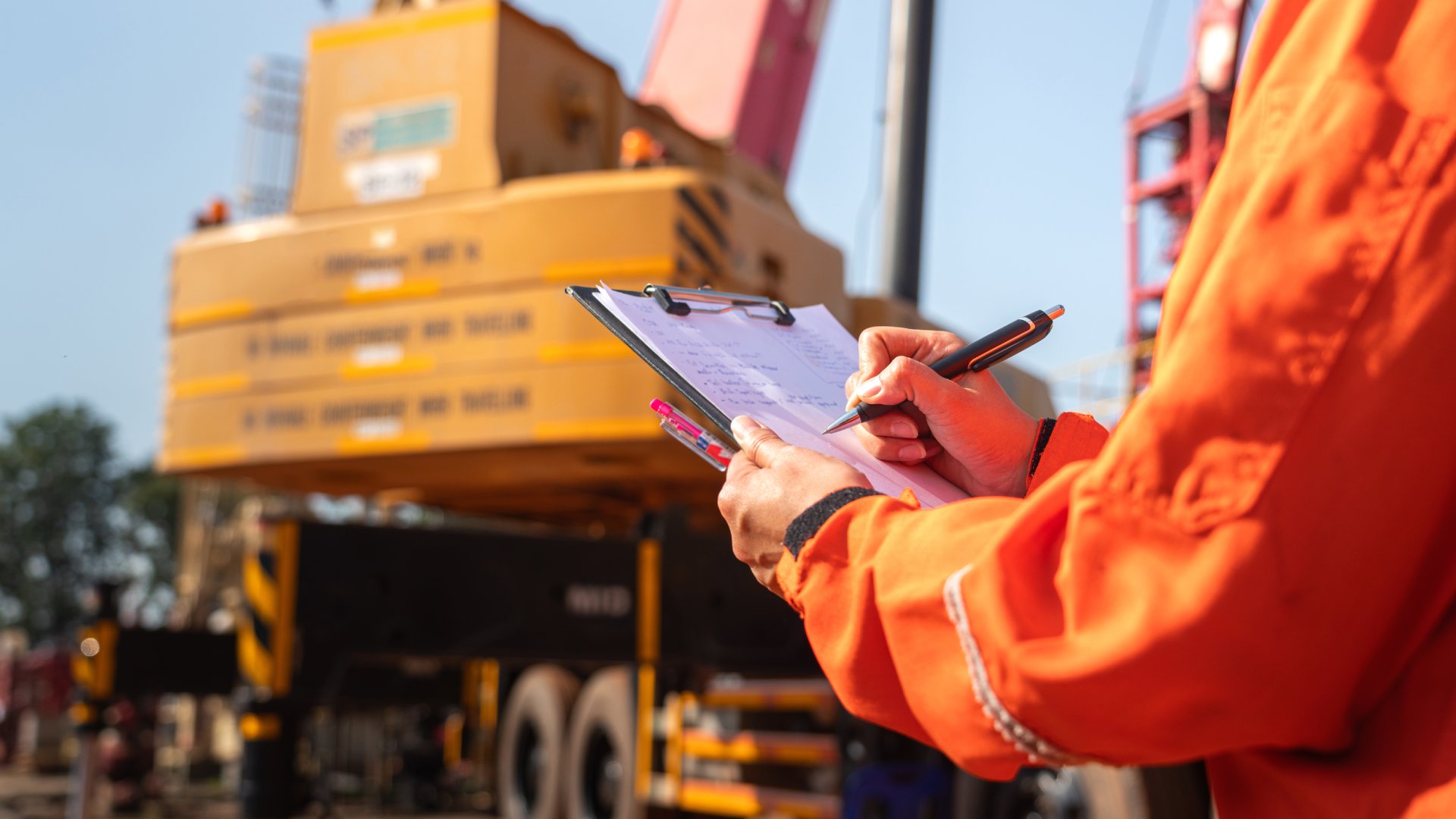 Close-up of a person wearing an orange suit and holding a clipboard in front of a large piece of heavy equipment.