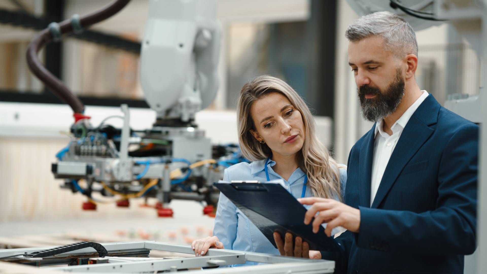 A man in a suit and a woman in a work shirt stand next to each other, looking at a clipboard with machines behind them.