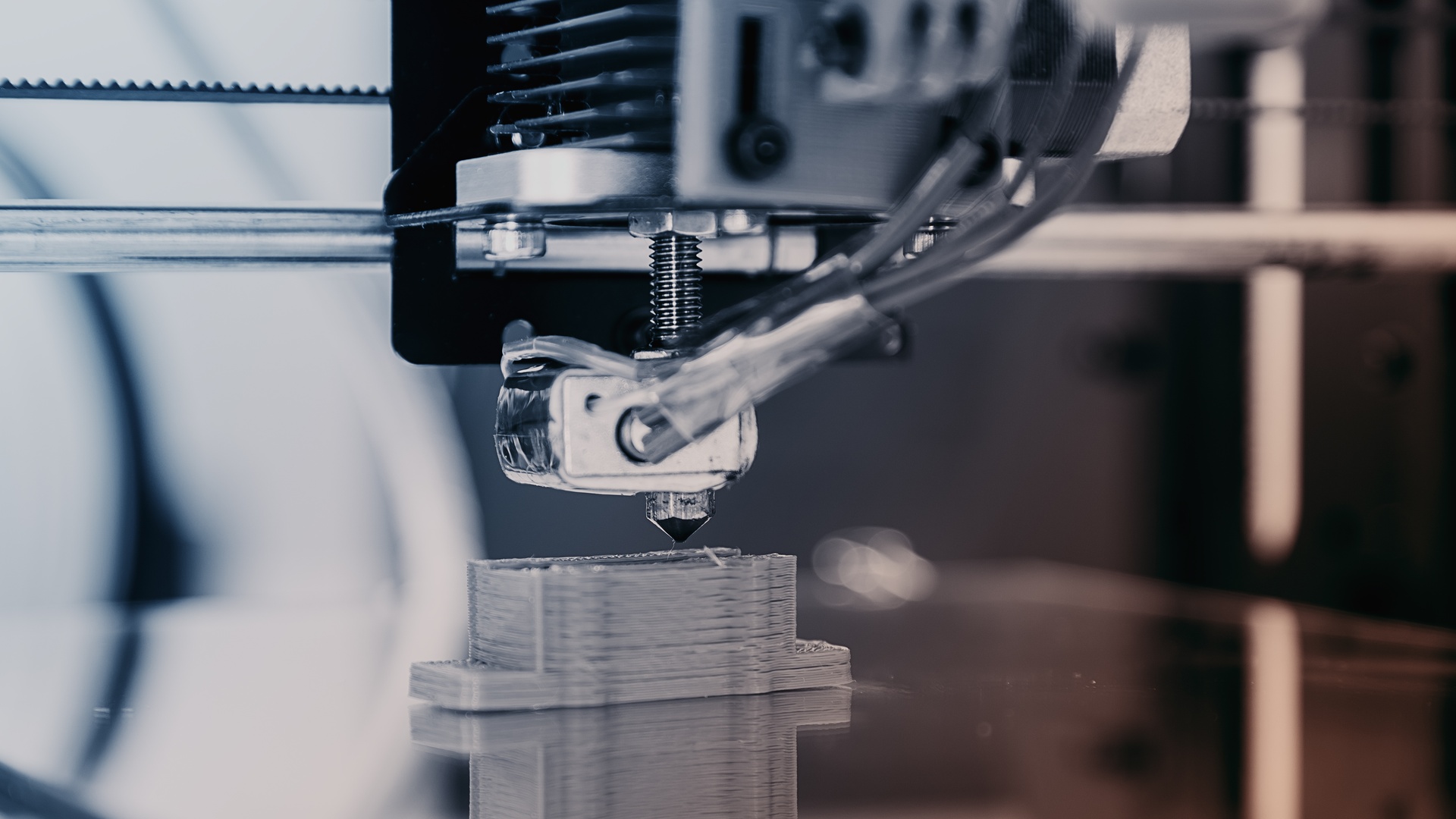 A close-up of a 3D printing machine producing a small gray rectangular plastic component with a wider base.