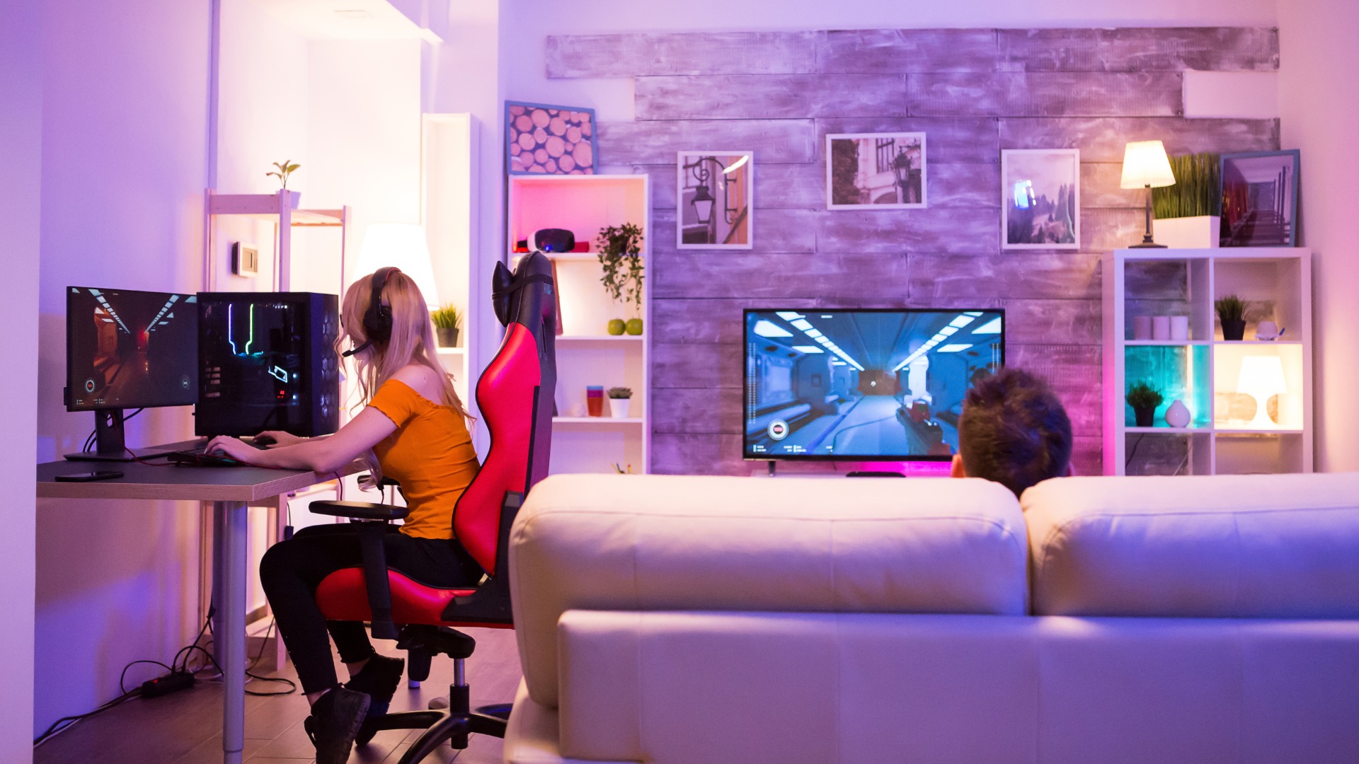 A young woman in a home game room plays games on a computer. Another person sits on a couch and plays on a TV.