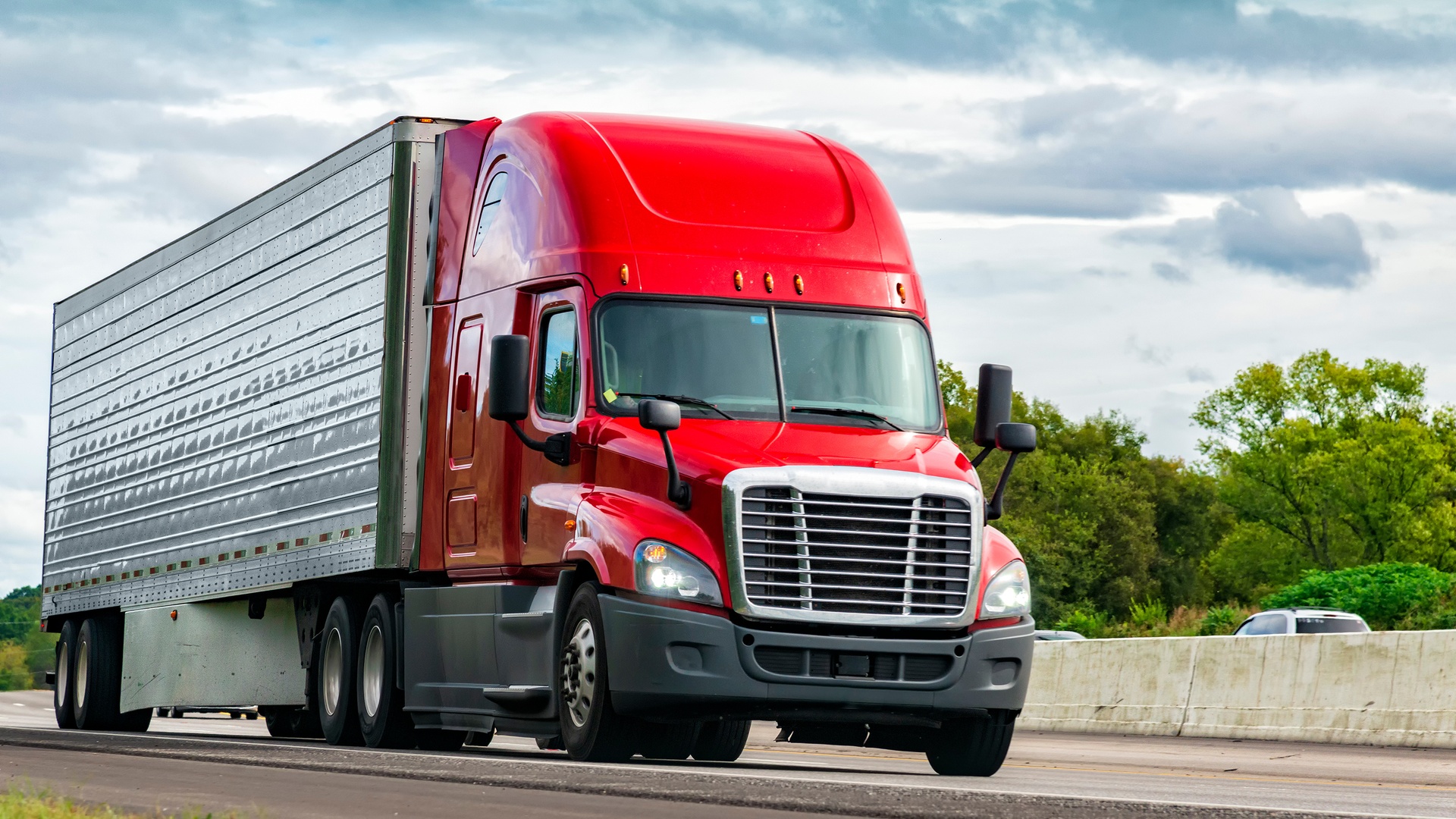 A red semi-truck with a trailer attached traveling down the right lane of an interstate highway under a cloudy sky.