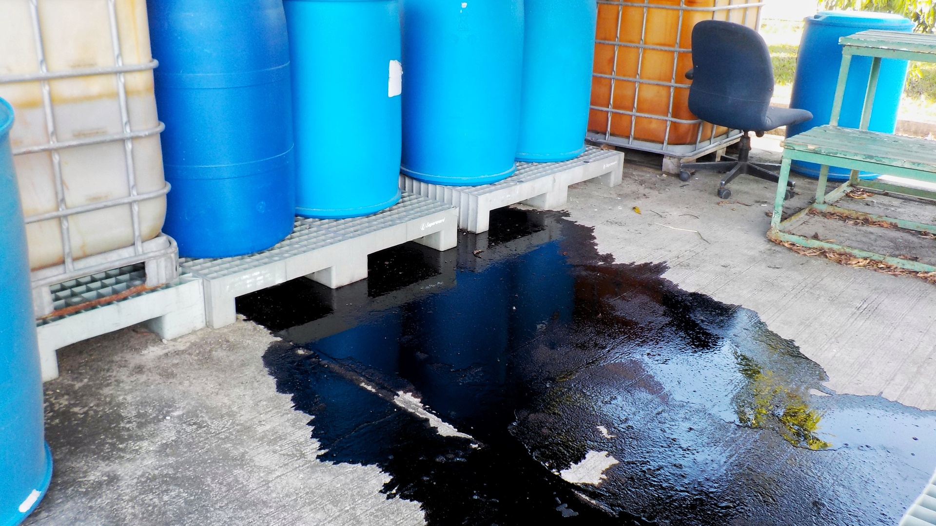 Blue drums stacked on top of pallets leak black liquid onto the concrete floor of an outdoor storage area.